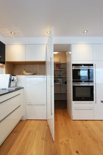 Moderne weiße Küche mit Holzboden, integrierte Backöfen und geöffnete Speisekammer.
