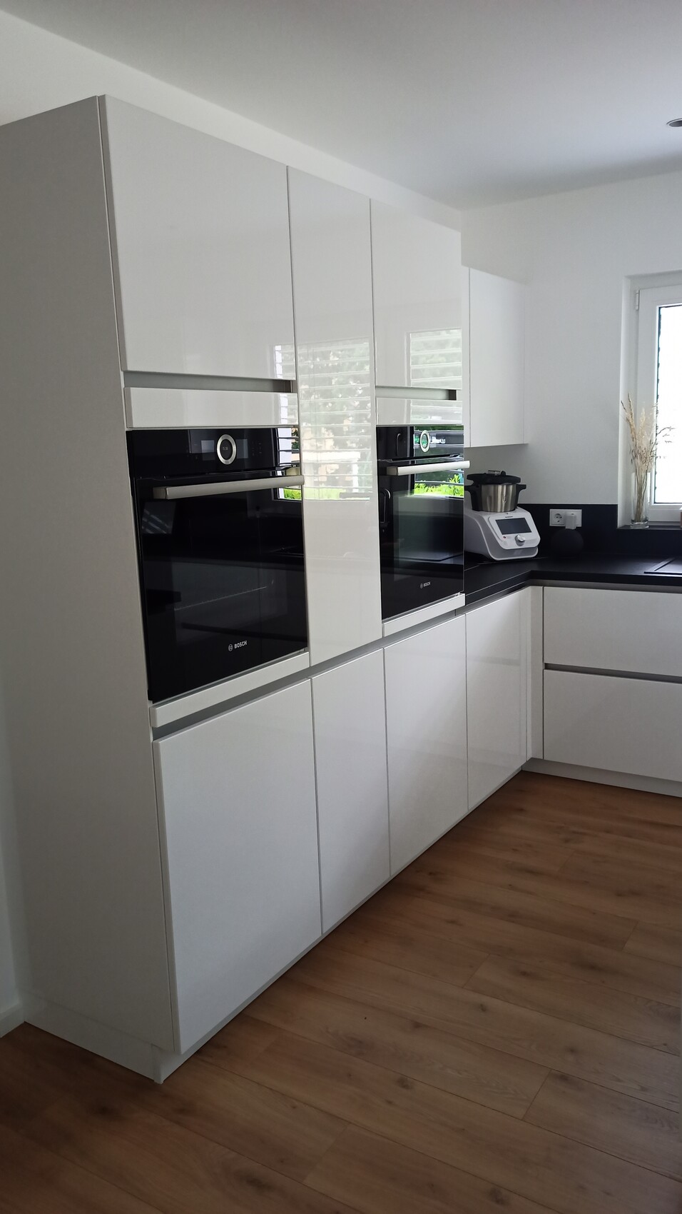 Moderne Küche mit weißen Hochglanz-Schränken, integrierten Backöfen und dunkler Arbeitsfläche.