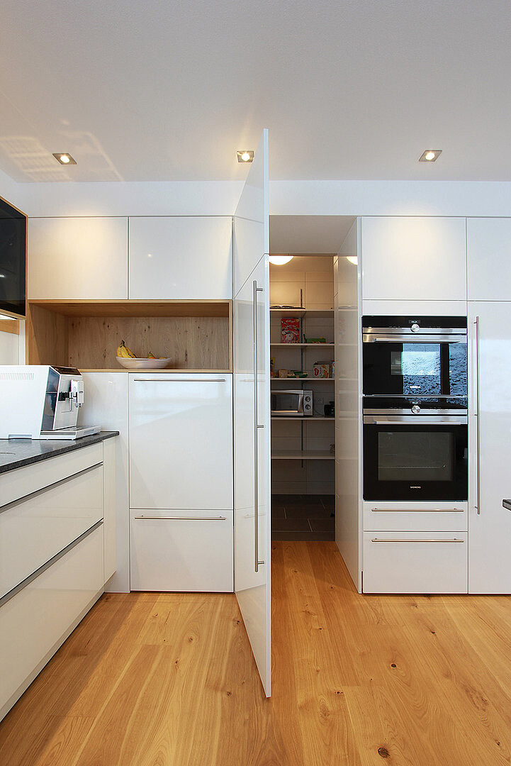 Moderne weiße Küche mit Holzboden, integrierten Backöfen und offener Vorratskammer dahinter.
