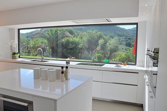 Moderne weiße Küche mit Insel, großer Fensterfront, Blick auf Garten mit Palmen.