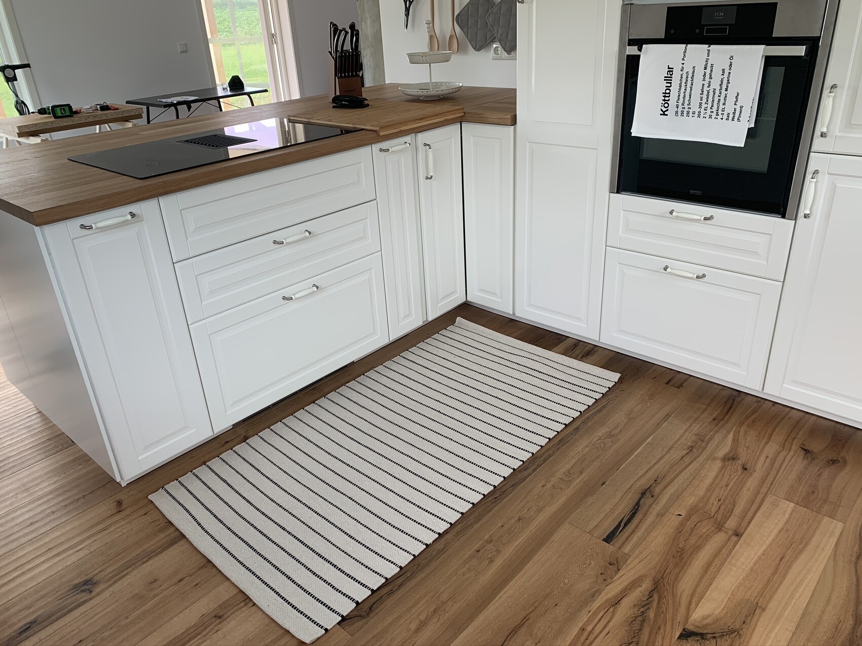 Moderne weiße Küche mit Holzarbeitsplatte, Kochinsel, Backofen und gestreiftem Teppich auf Holzboden.