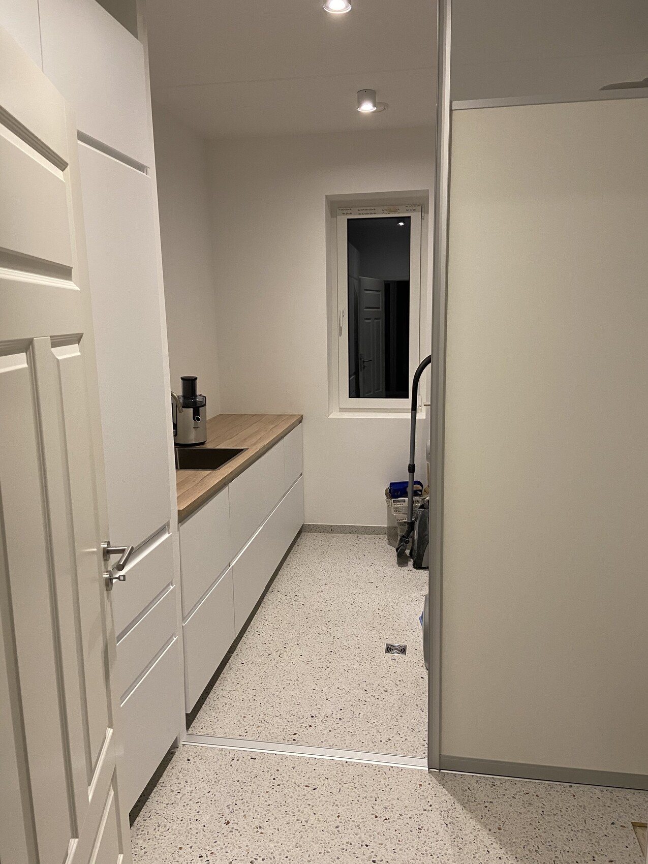 Küche: weiße Schränke, Holzarbeitsplatte, Spüle, Küchengerät, Staubsauger am Fenster.
