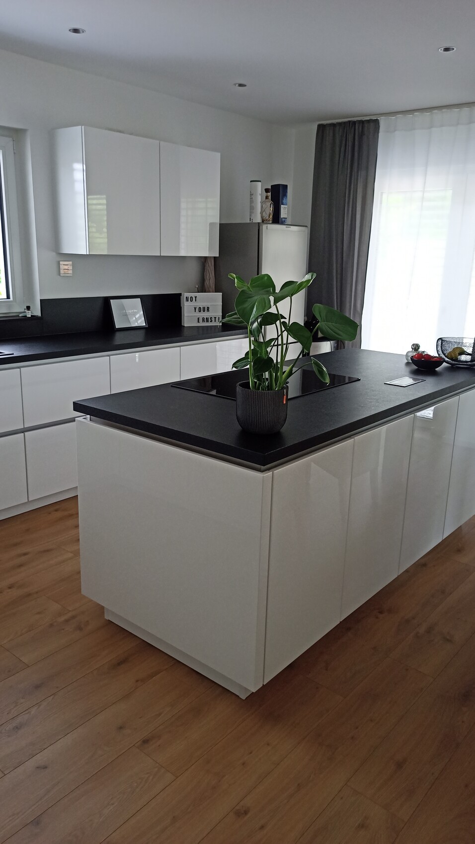 Moderne Küche: weiße Schränke, schwarze Insel, Pflanze, Kühlschrank, Vorhänge.