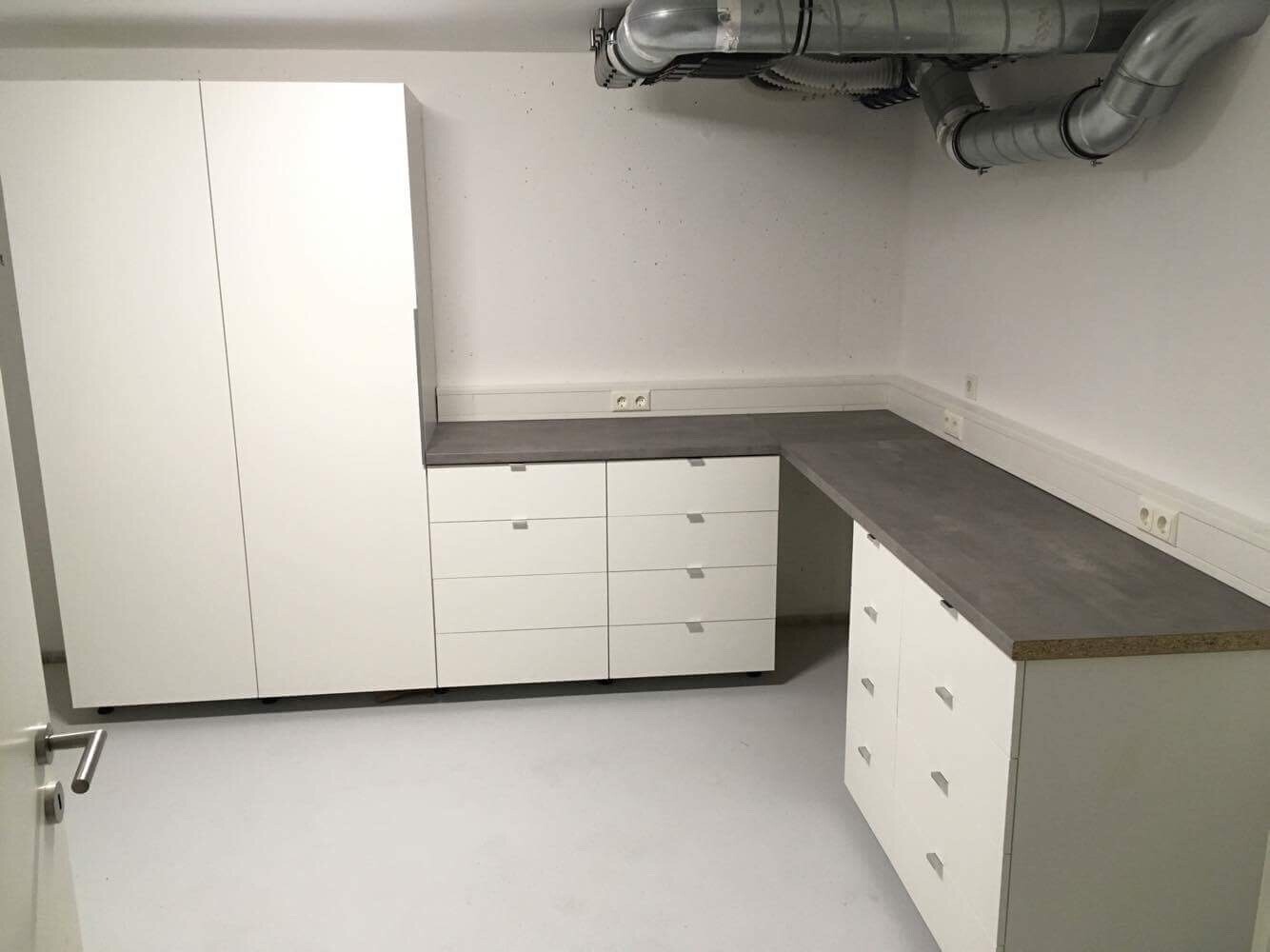 Küche mit weißen Schränken, grauer Arbeitsplatte und L-Form; sichtbare Lüftungsrohre an der Decke.