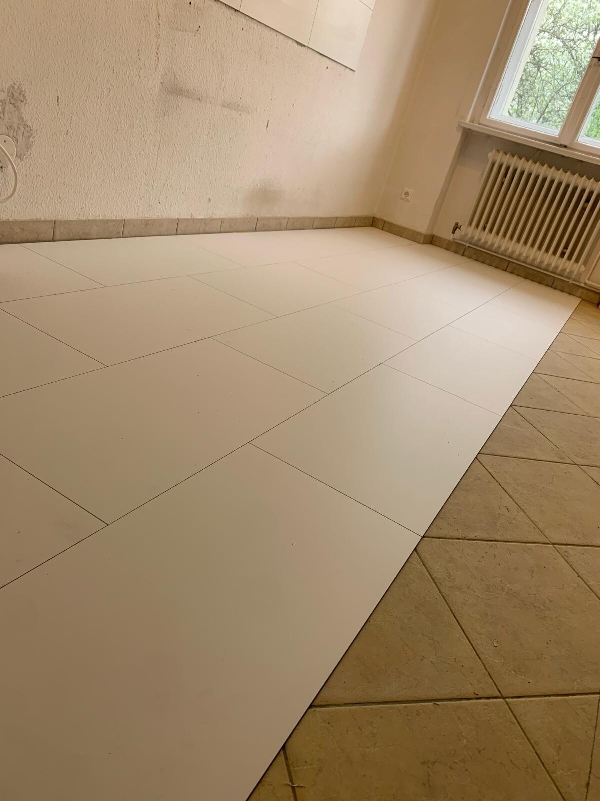 Leeres Zimmer mit weißen Großfliesen, Fenster und Heizkörper.