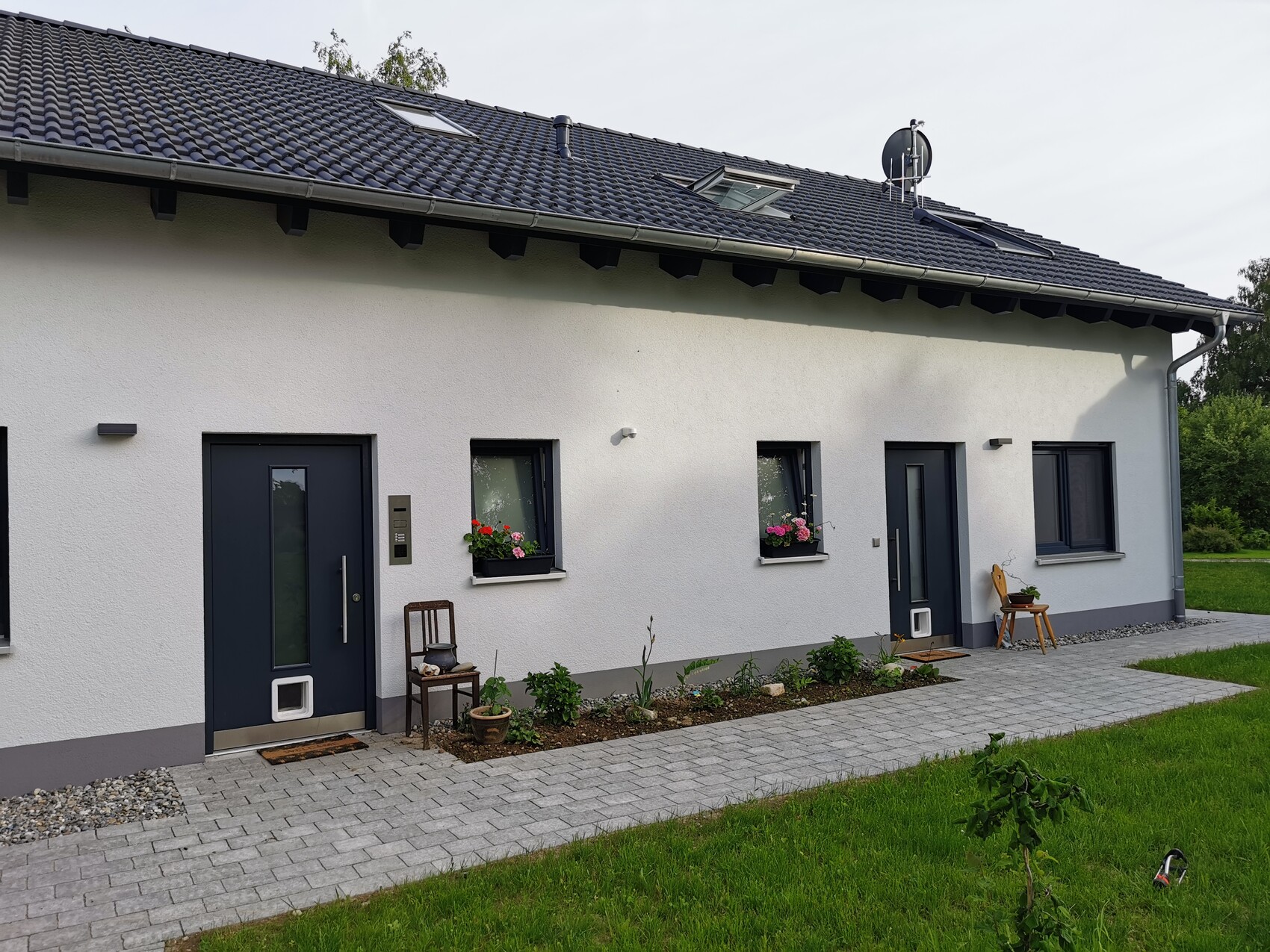 Weiße Wohnhausfront mit zwei dunkelblauen Türen, Fenstern mit Blumenkästen und gepflastertem Weg.