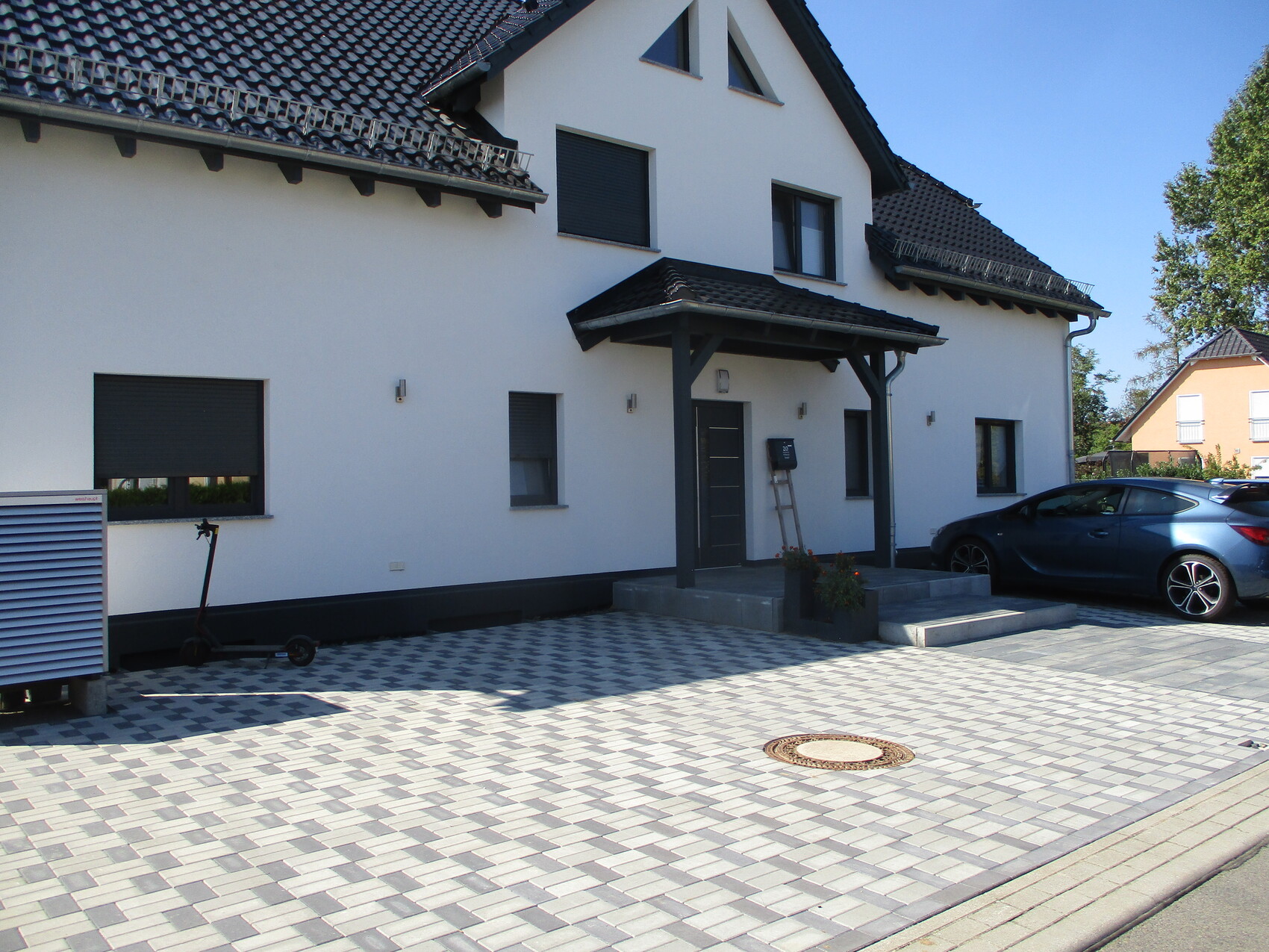 Weißes Einfamilienhaus mit dunklem Dach, gepflasterter Vorplatz, geparktem Auto und Scooter.