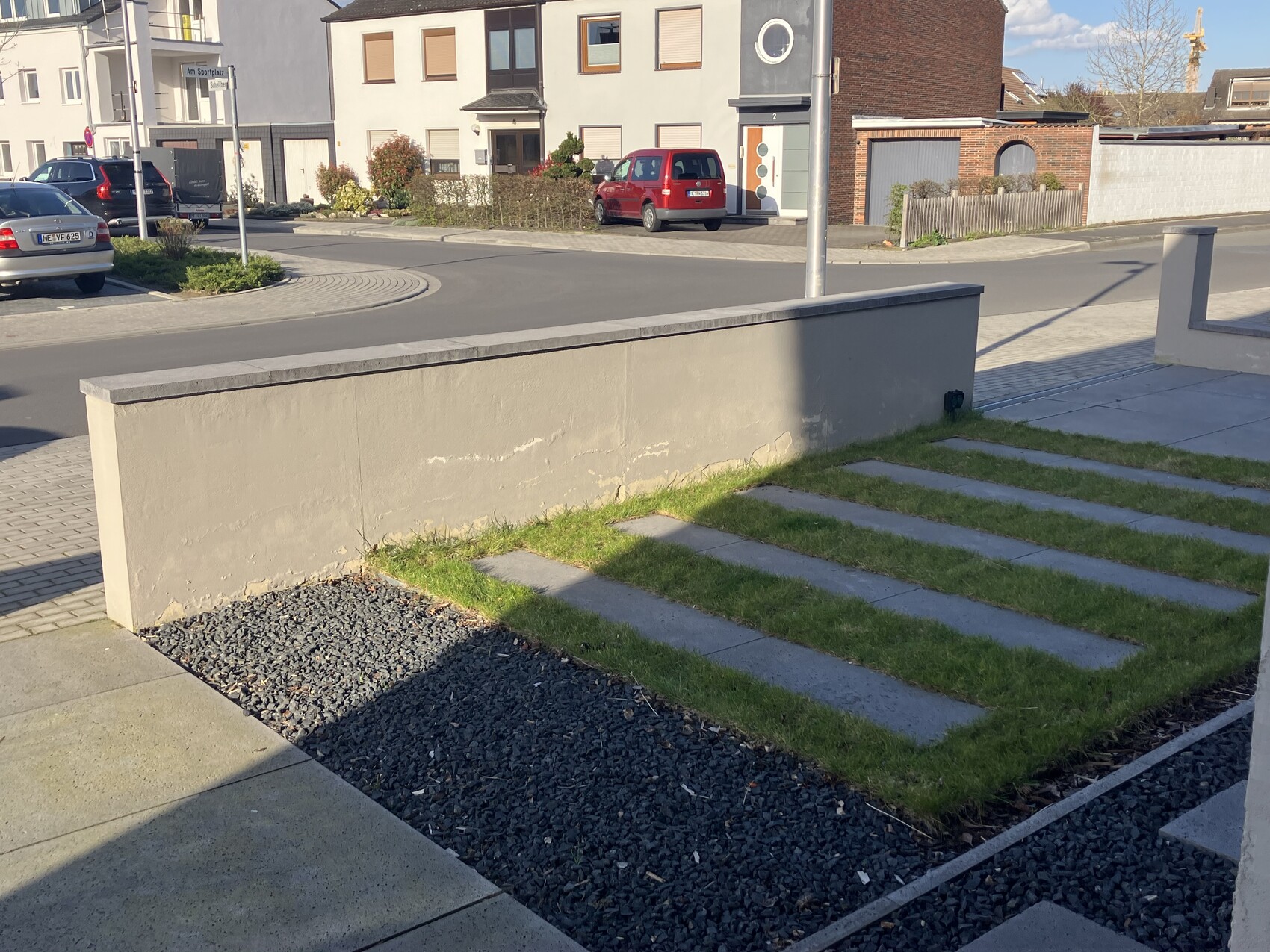 Frontansicht eines Vorgartens mit Betonmauer, Grasstreifen und Steinplatten, Straße im Hintergrund.