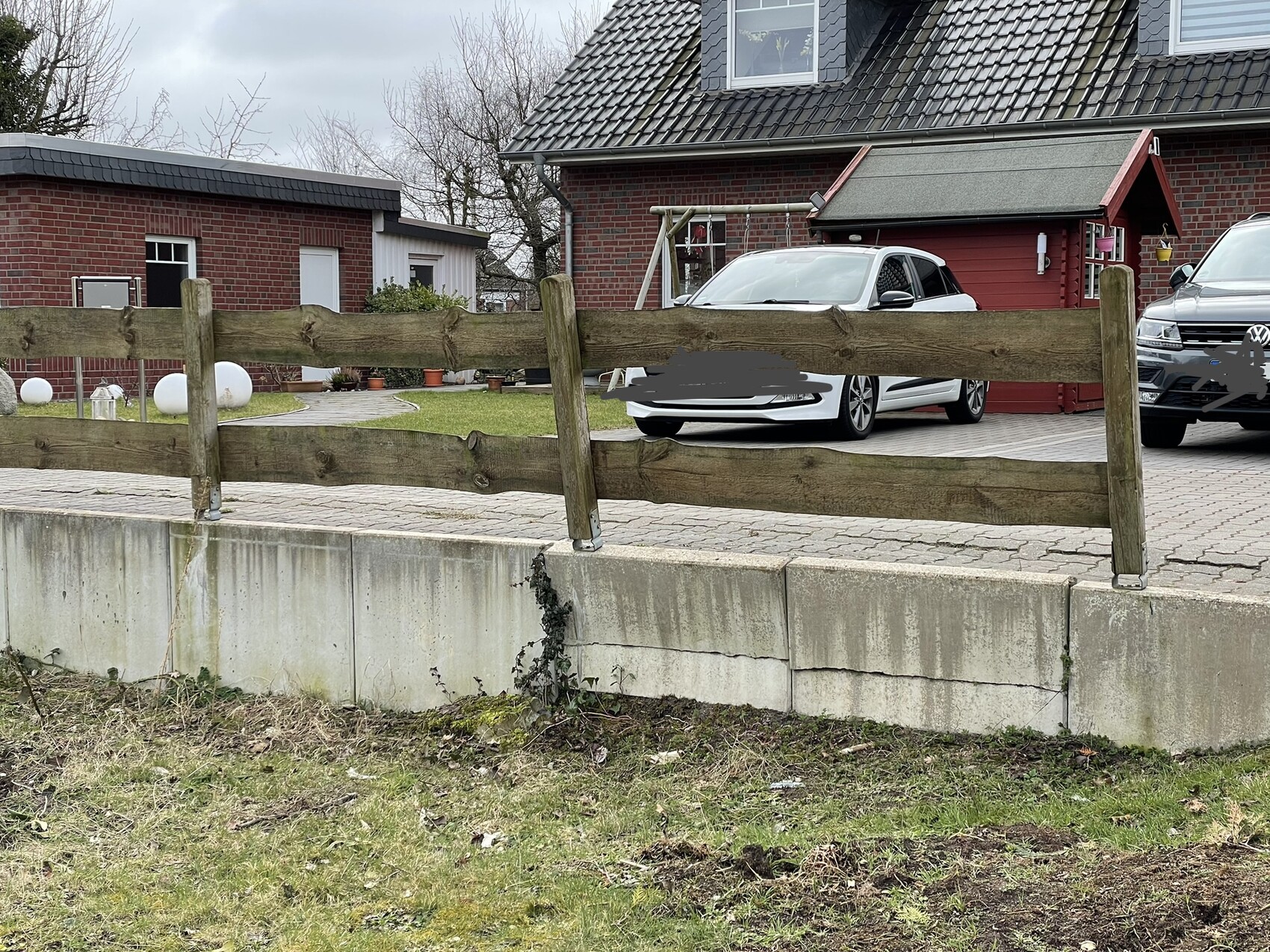 Holzlattenzaun vor Betonmauer, dahinter weißer PKW und grauer SUV vor rotem Haus.