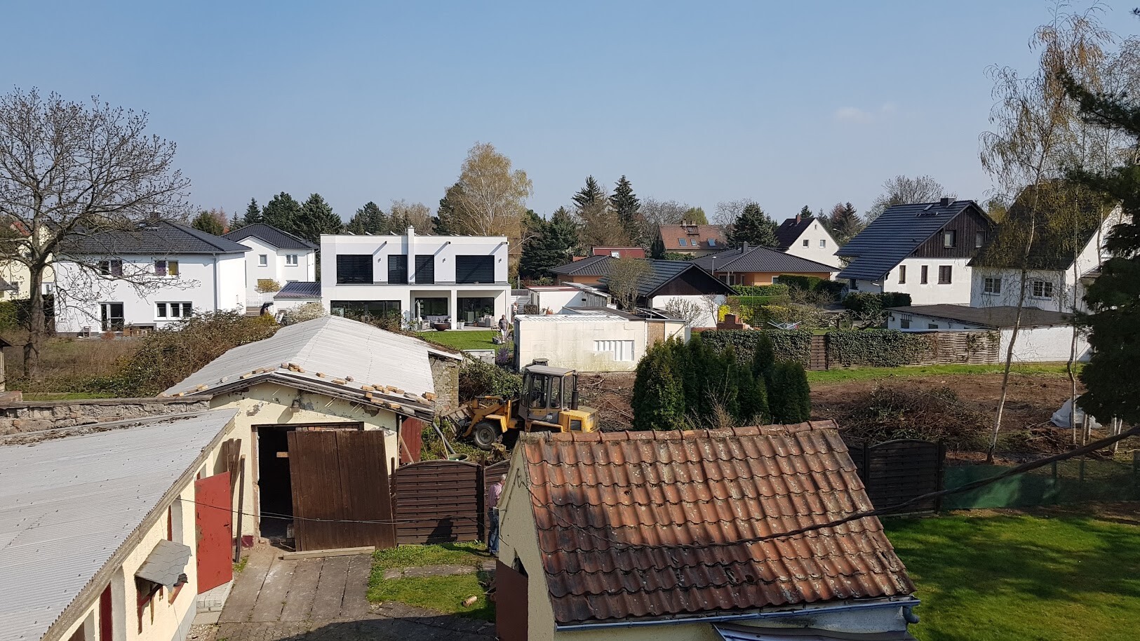Vorstadt mit modernen weißen Häusern; gelber Bagger auf Baufläche, Bäume im Hintergrund.