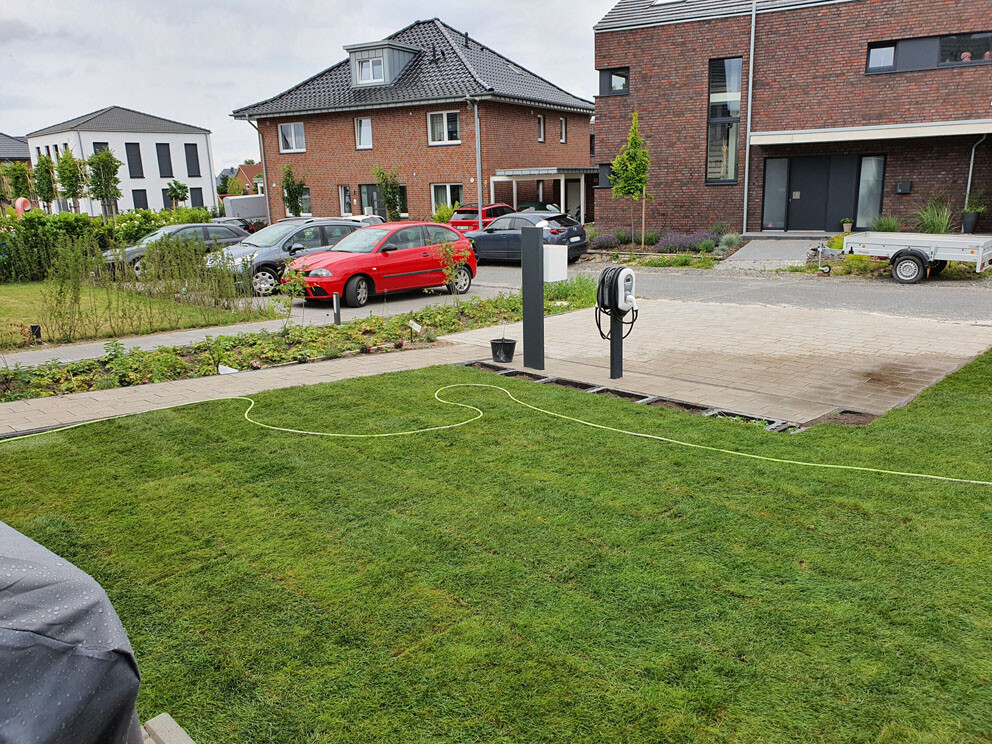 Vorstadtgarten mit Zufahrt, Schlauchaufroller und geparkten Autos vor Backsteinhäusern.