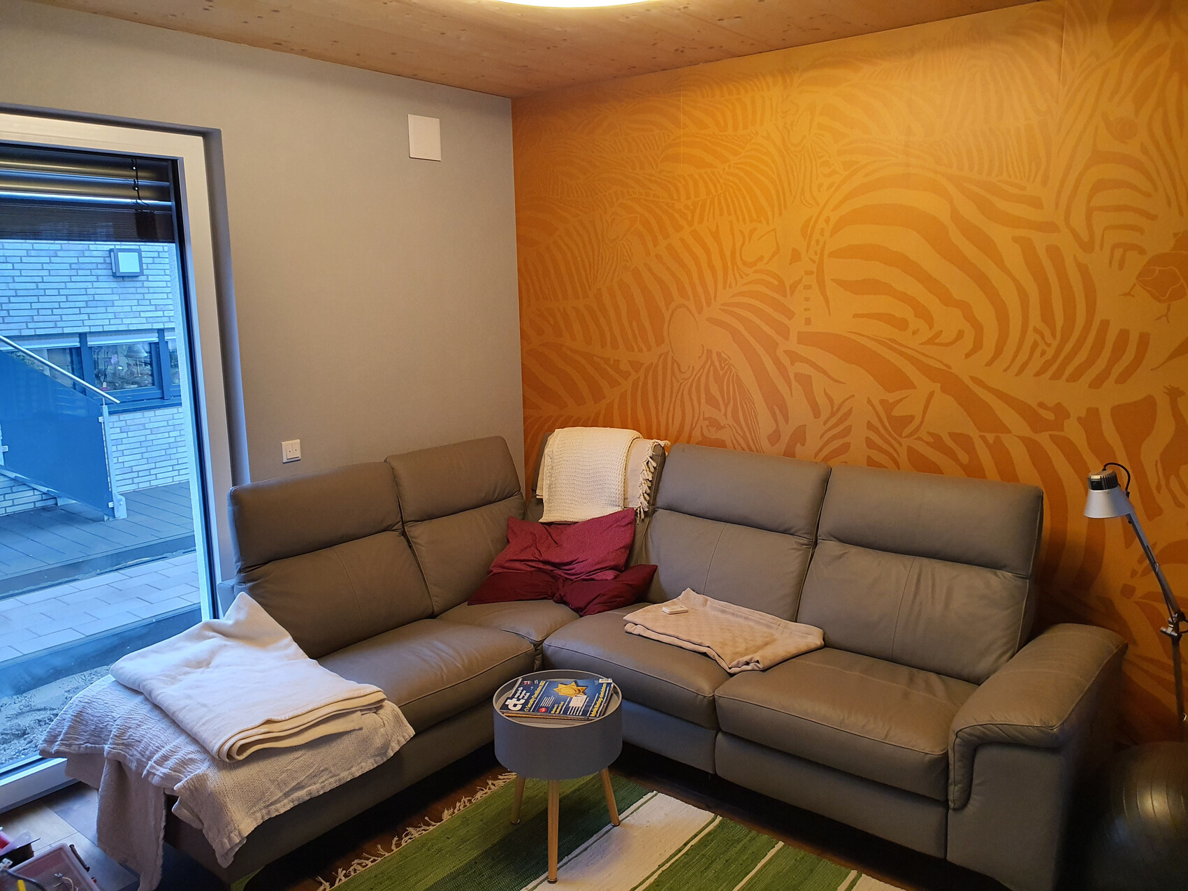 Wohnzimmer mit grauer Eckcouch, orangener Zebra-Musterwand, Fenster zum Balkon, Decken und Kissen.