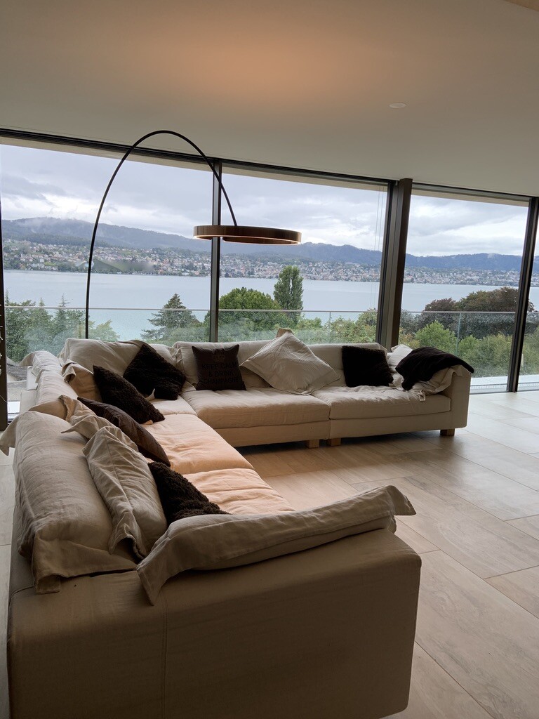 Großes beiges Ecksofa mit vielen Kissen im Wohnzimmer vor Fensterfront mit Seeblick und Berge.