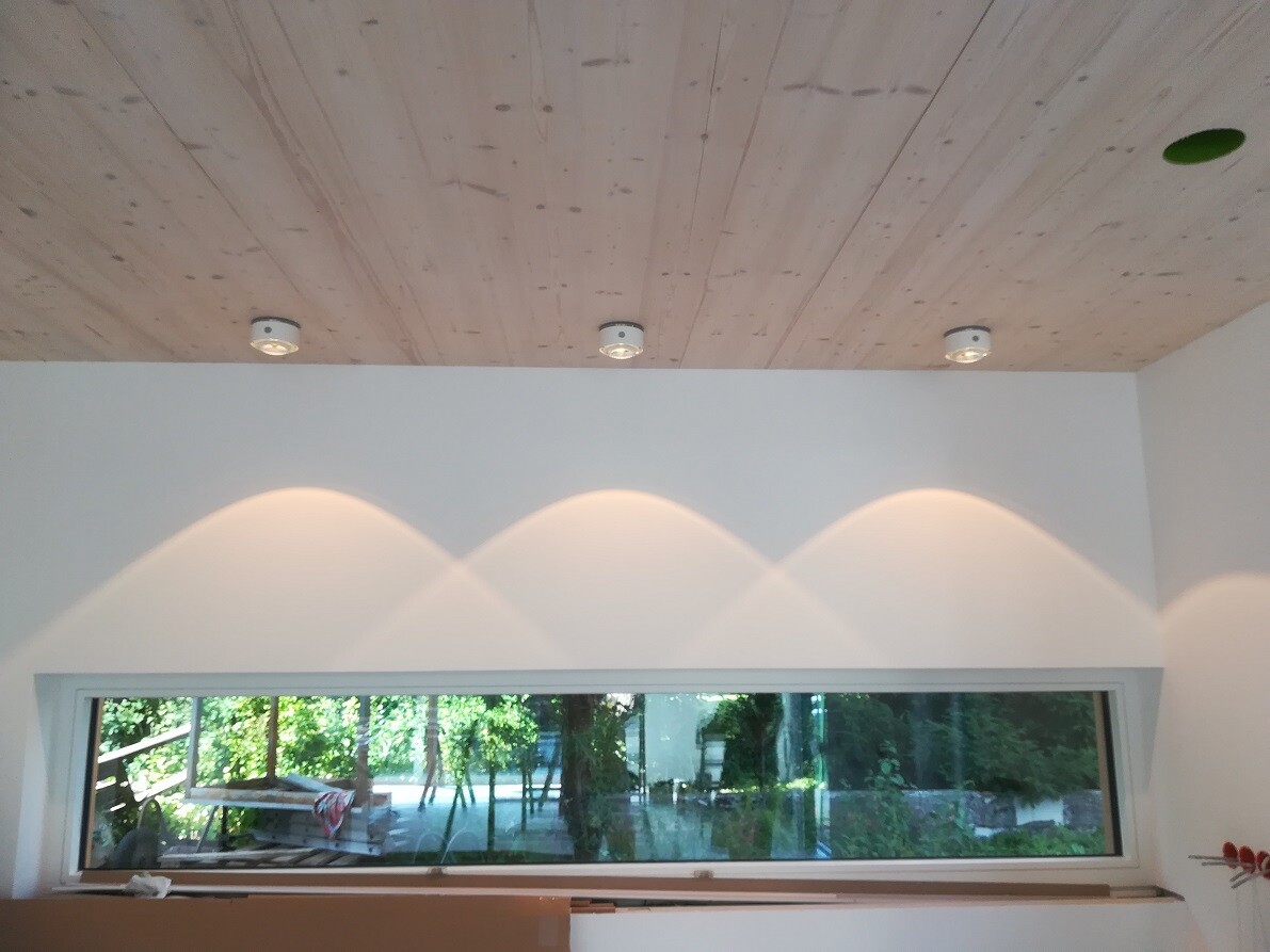 Heller Innenraum mit Holzbalkendecke, drei Deckenspots und langem horizontalem Fenster, Gartenblick.