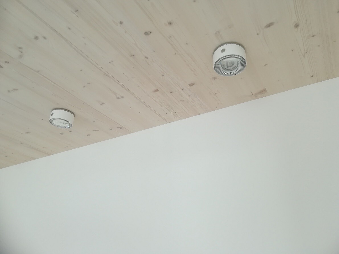 Holzdecke mit zwei runden weißen Deckenlampen über einer hellen Wand.