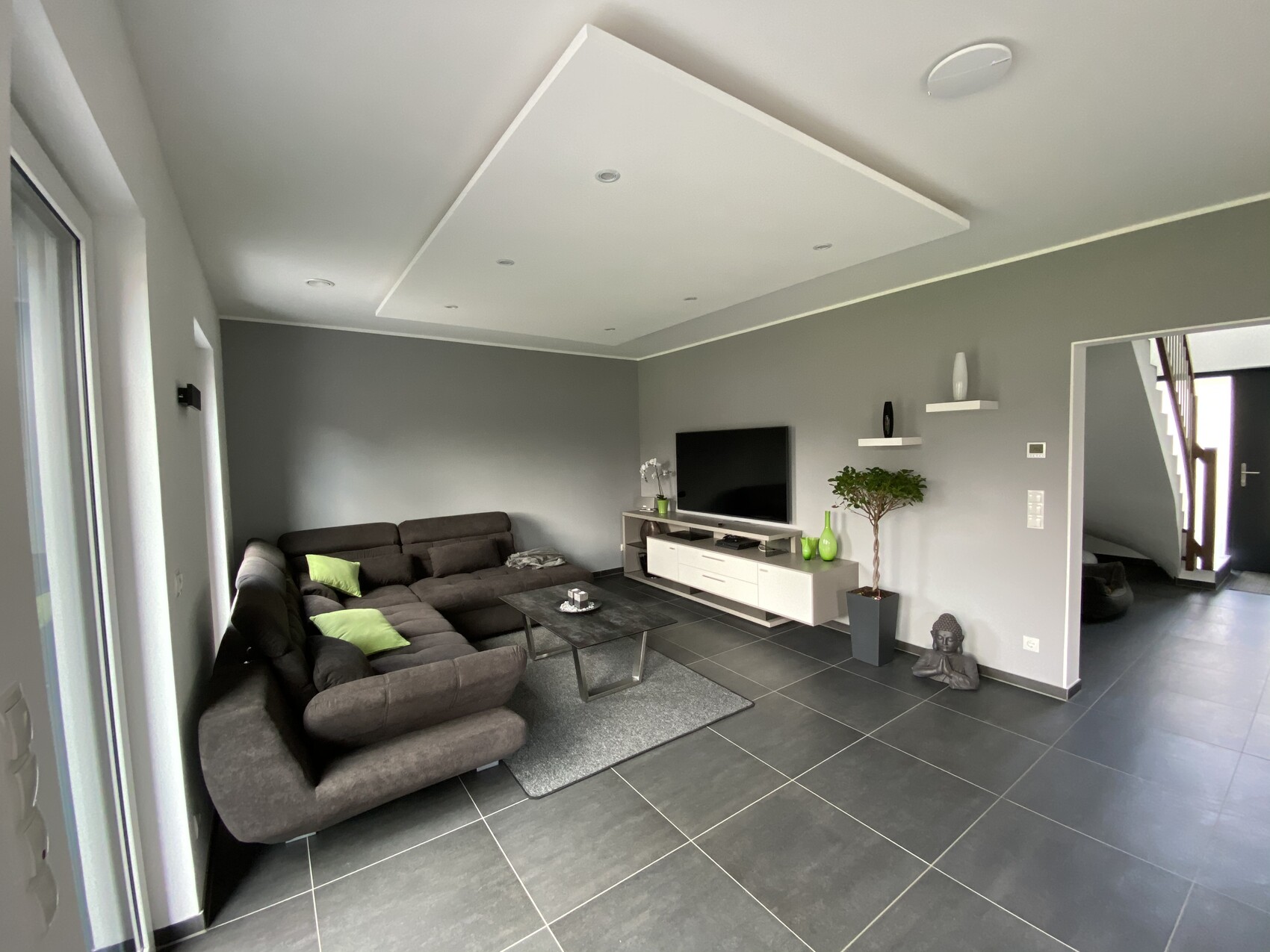 Modernes Wohnzimmer: graue Wände, dunkles Ecksofa, TV-Mediestation, Pflanze und Buddha-Statue.