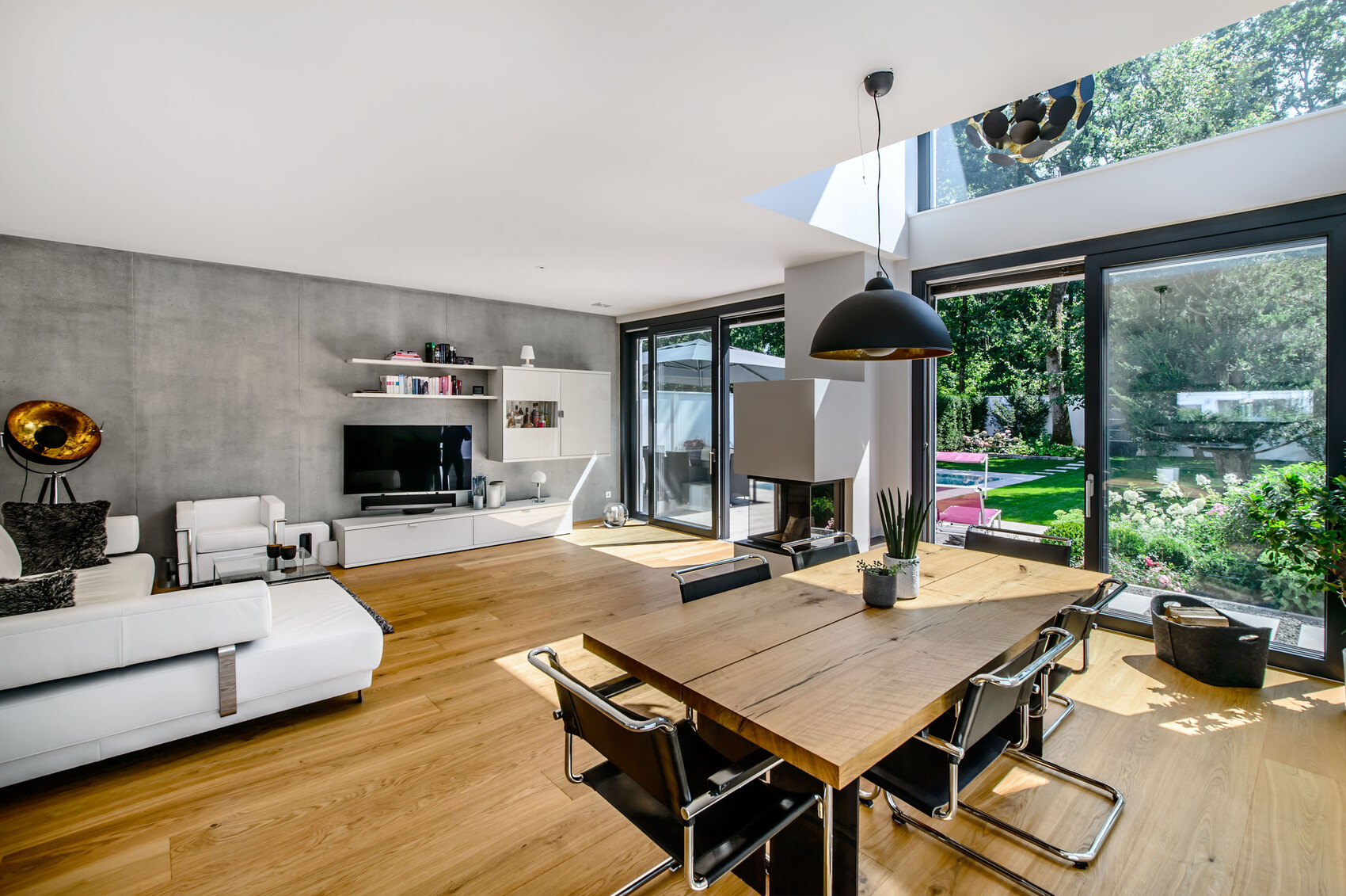 Offenes Wohn- und Esszimmer mit weißer Couch, Holztisch und großen Glasfenstern zum Garten.