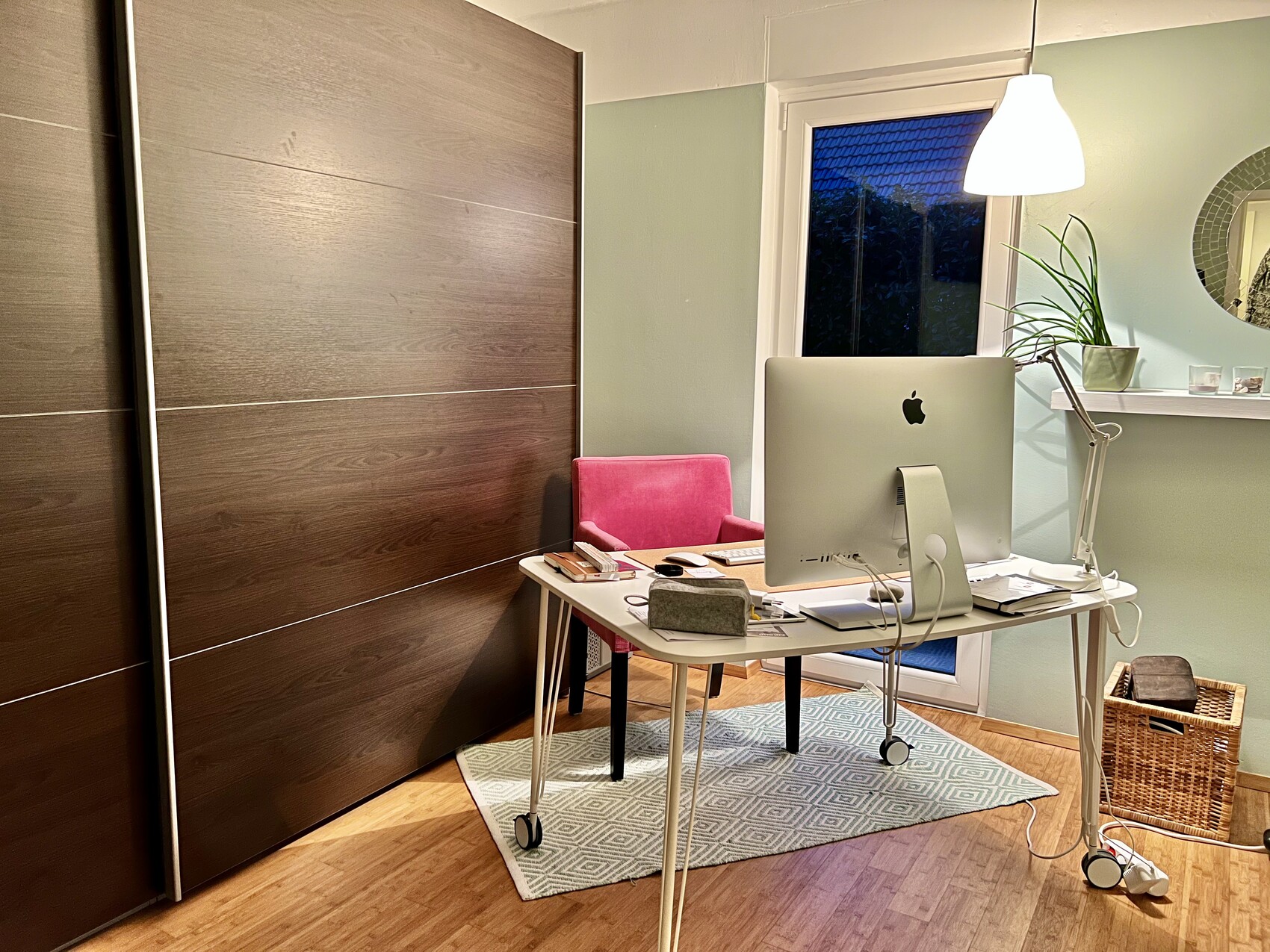 Arbeitszimmer mit iMac auf Tisch, pinkem Stuhl, Pflanze, Fenster mit Abendblick und braunem Schrank.