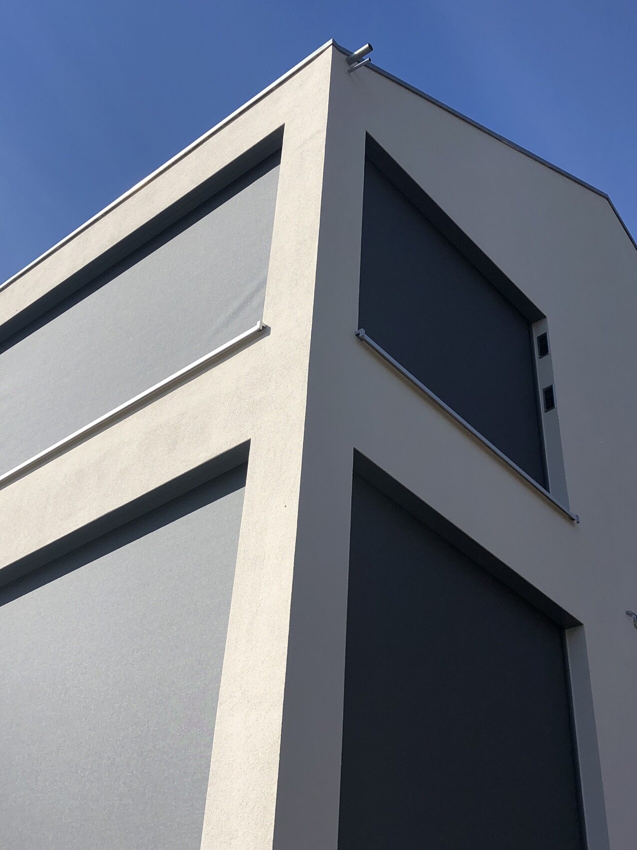 Moderne weiße Gebäudeecke mit zwei großen, geschlossenen dunklen Rollläden vor Fenstern; blauer Himmel.