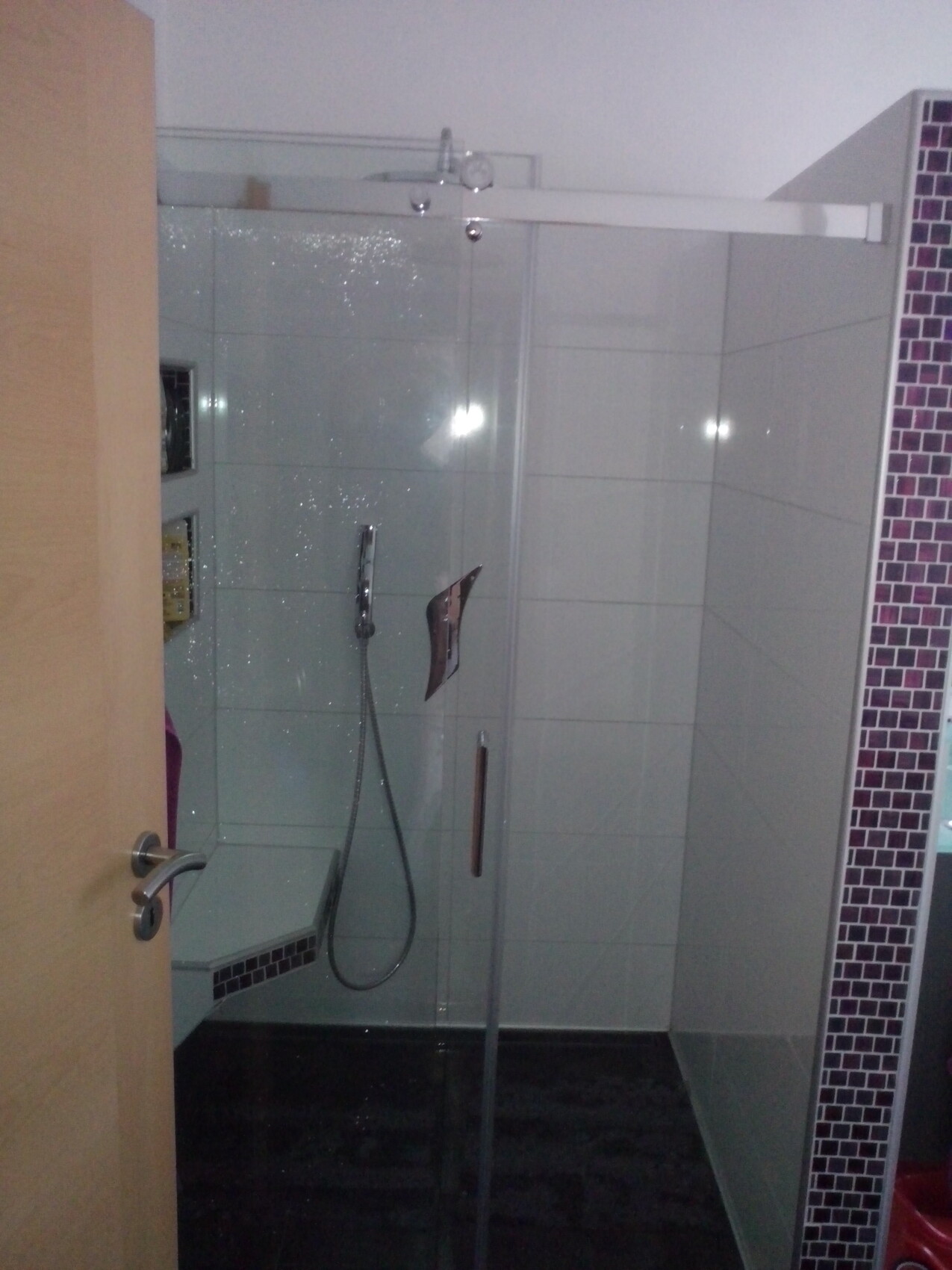 Offene Tür führt zu einer Glas-Duschkabine mit Handbrause, weiße Fliesen, violette Mosaikwand.