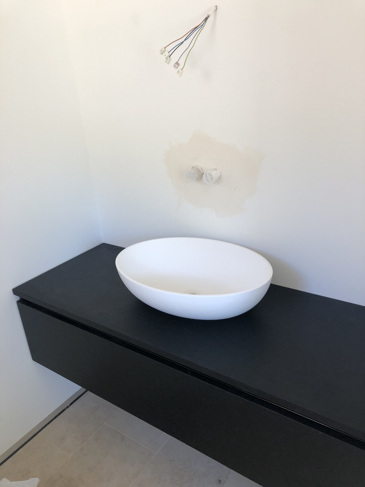 Weiße runde Waschschüssel auf schwarzer Konsole; lose Kabel hängen an der Wand.