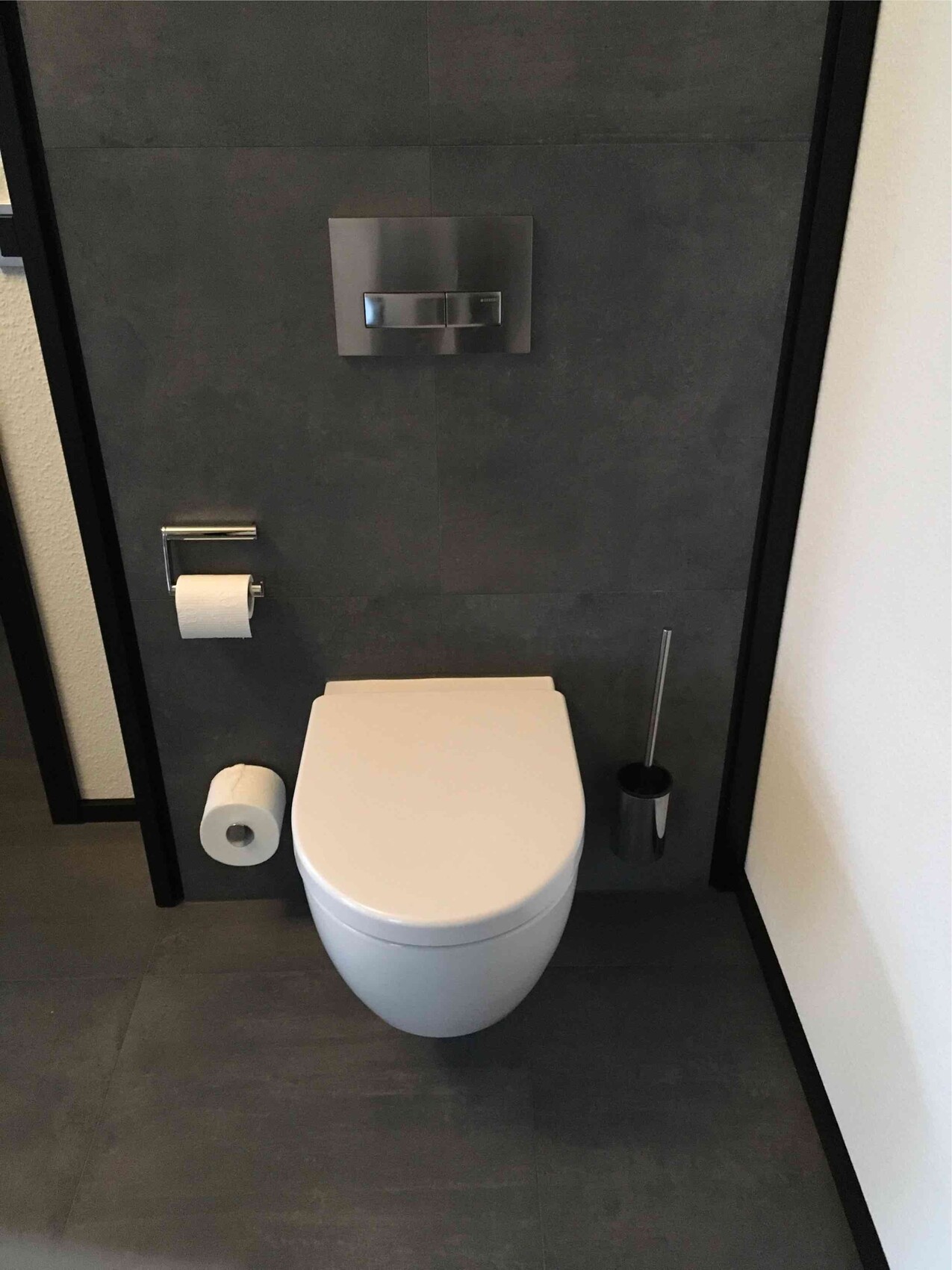 Moderne Toilette vor dunklen Fliesen mit Wandspülkasten; links Papierrolle, rechts Bürste.