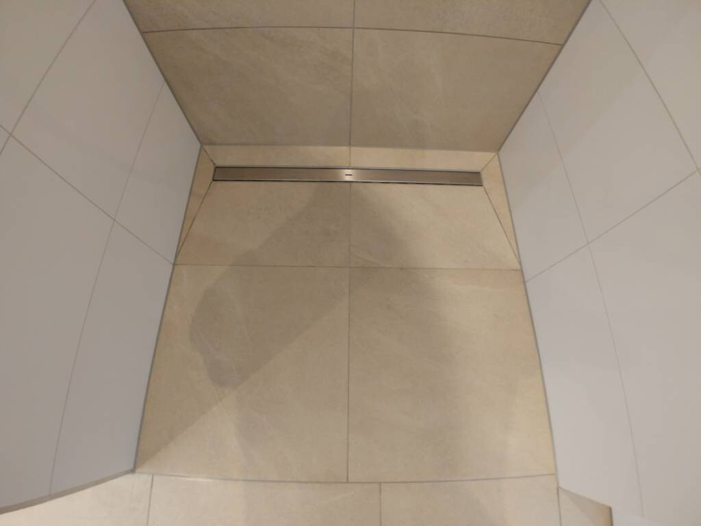 Duschboden mit linearer Ablaufrinne aus Edelstahl, beige Fliesen und weiße Wandfliesen.