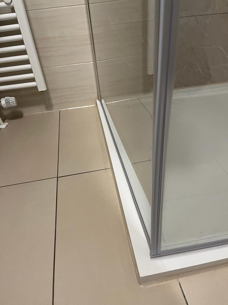 Eckbereich einer Dusche mit Glasschiebetür, weißem Rahmen und hellen Fliesen.