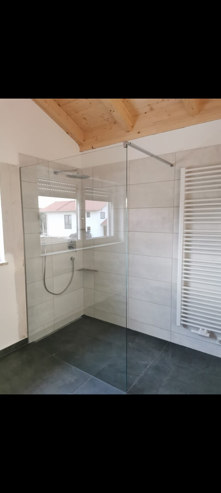 Offene Glasdusche mit grauen Wand- und Bodenfliesen, Fensterblick, Handbrause, Holzdecke.