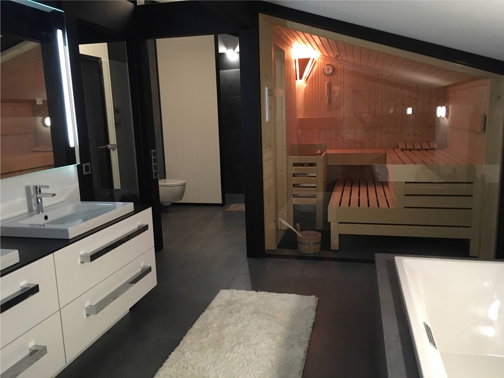 Modernes Badezimmer mit Waschtisch, Toilette und offener Holzsauna hinter Glaswand