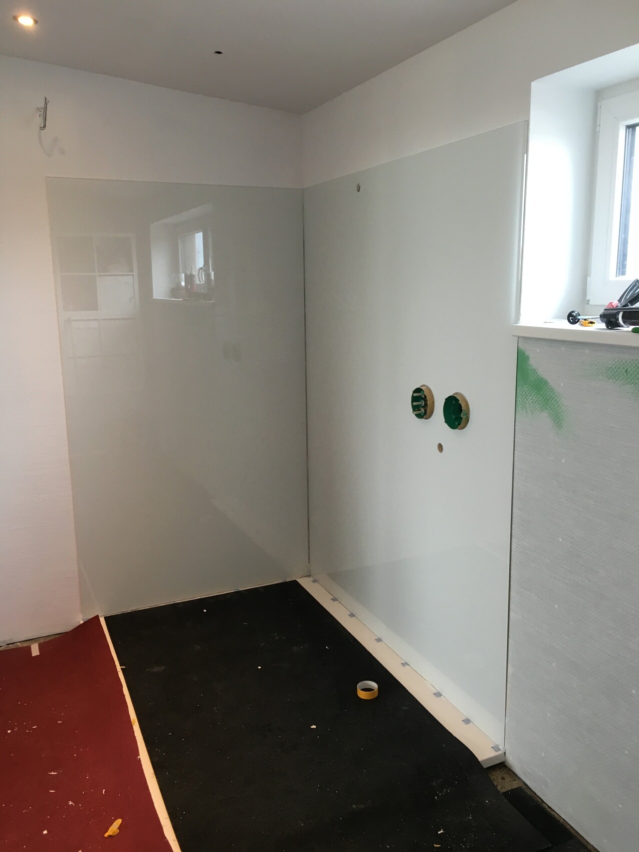 Badezimmerunterbau mit Glaswand-Dusche, offenen Installationen und Fenster rechts.