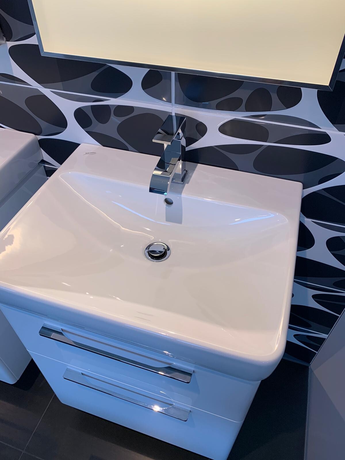 Modernes weißes Waschbecken mit Chromarmatur vor schwarz-weißen Wellenfliesen, Spiegelschrank.