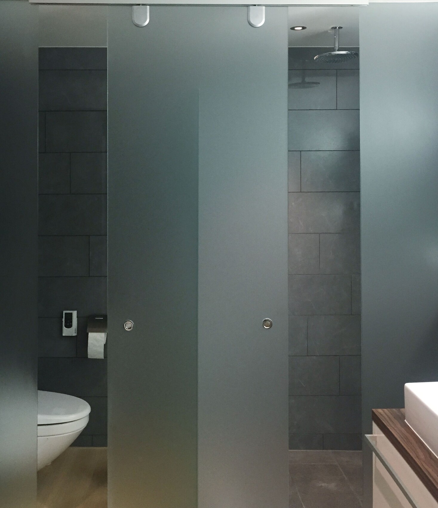 Moderne öffentliche Toilette mit grauen Trennwänden, dunklen Fliesen und Waschbecken.