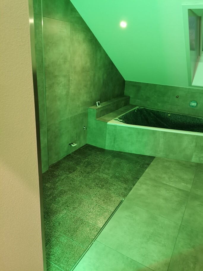 Grünes Badezimmer mit Eckbadewanne, dunklem Duschbereich und Smartphone auf der Ablage.