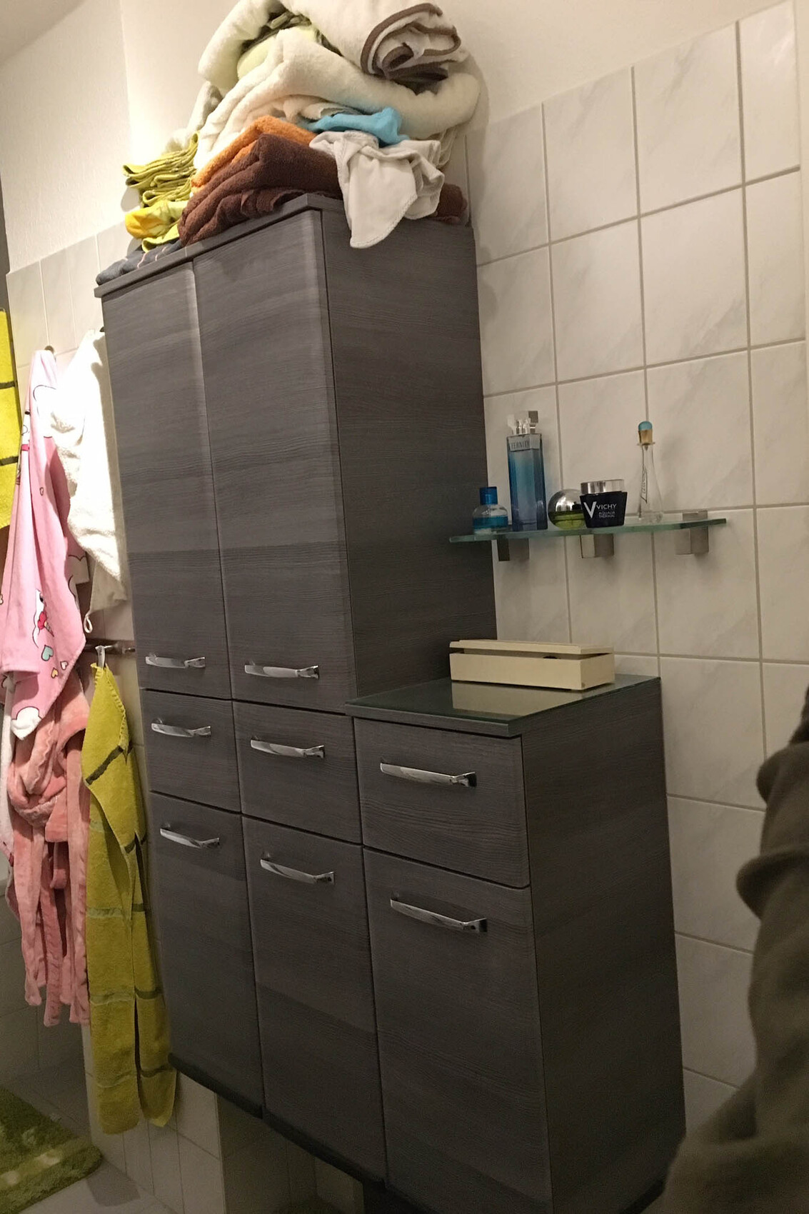 Modernes Badezimmer mit hohem grauen Badschrank, oben liegen Handtücher und Pflegeprodukte.