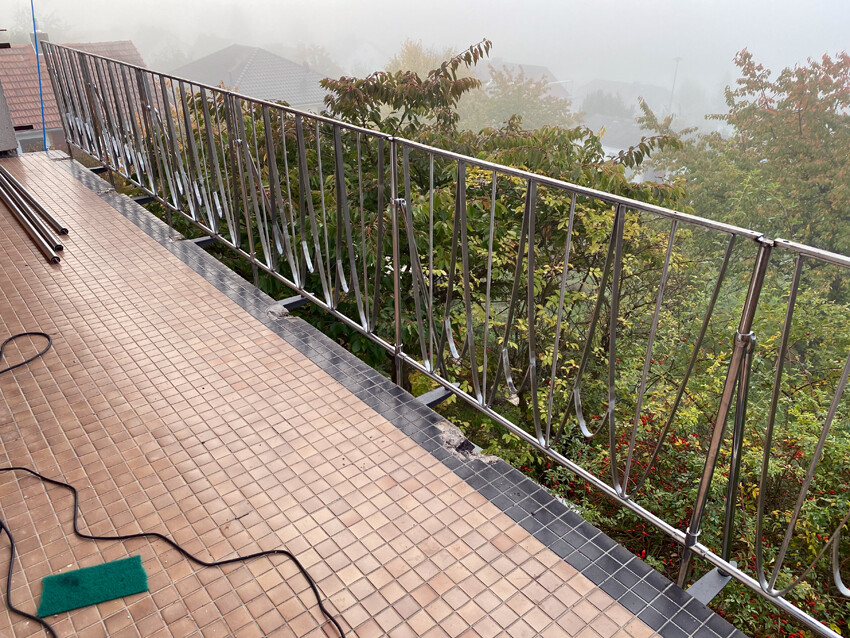 Balkon mit glänzendem Metallgeländer und beigen Fliesen; Nebel über den Bäumen.
