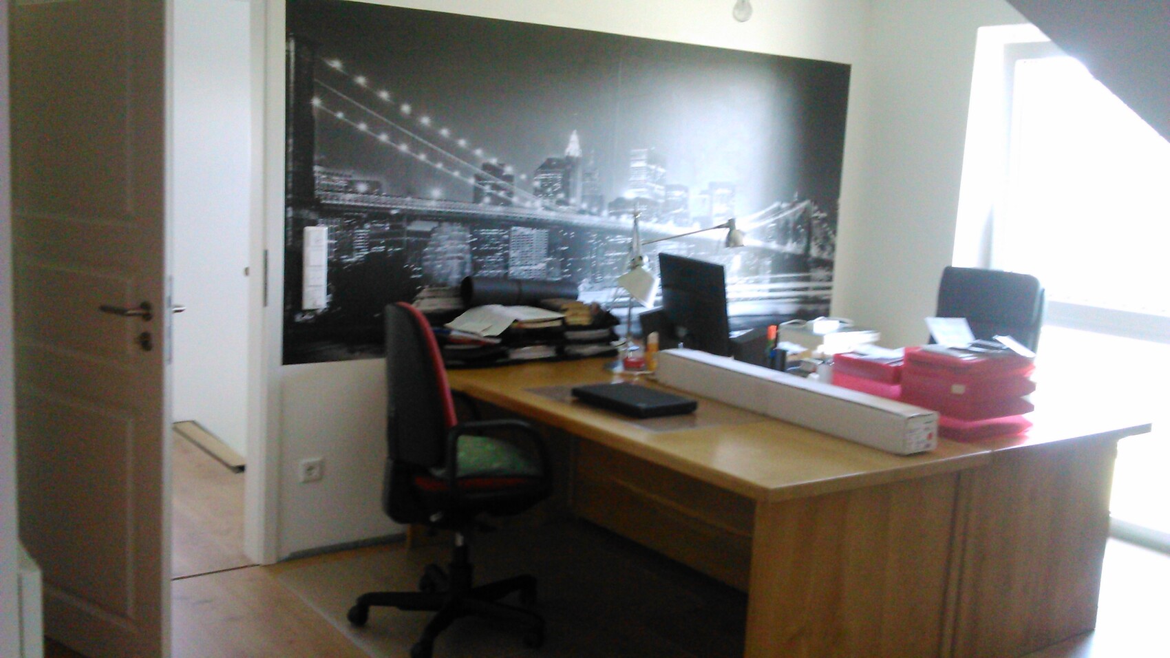 Büro-Schreibtisch mit Computer, Aktenstapeln und roten Ordnern vor einem Skyline-Wandbild der Brücke.