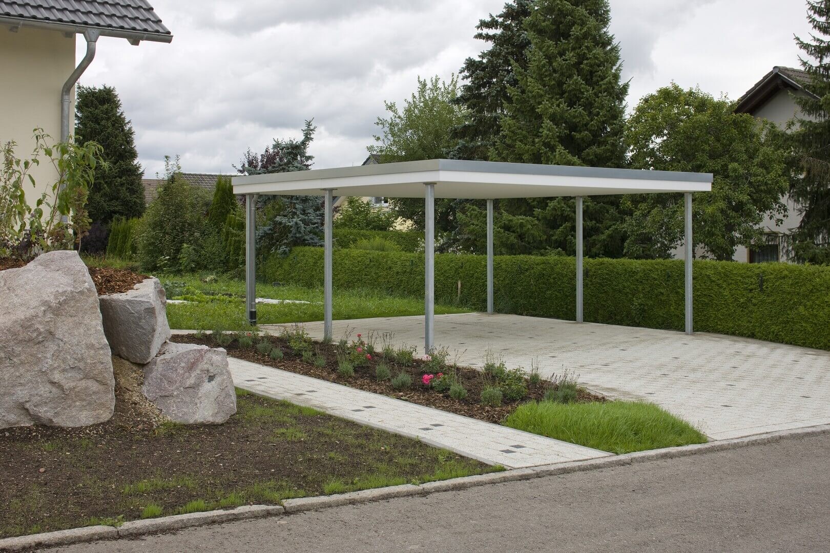 Gepflasterte Einfahrt mit weißem Carport aus Metall, von Büschen und Bäumen umgeben.