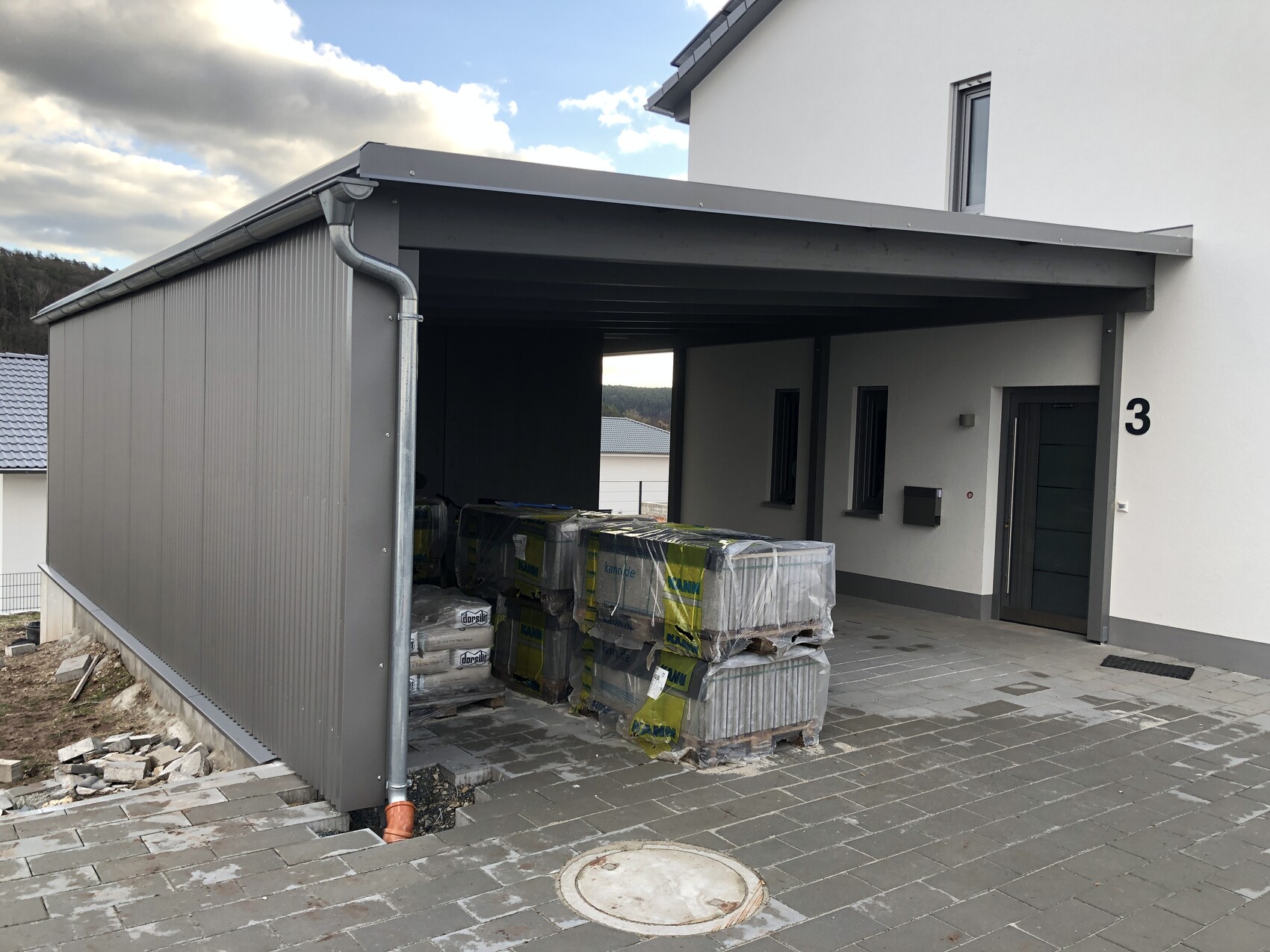 Außenansicht eines modernen Hauses mit grauem Metall-Carport; Baupaletten vor dem Eingang.