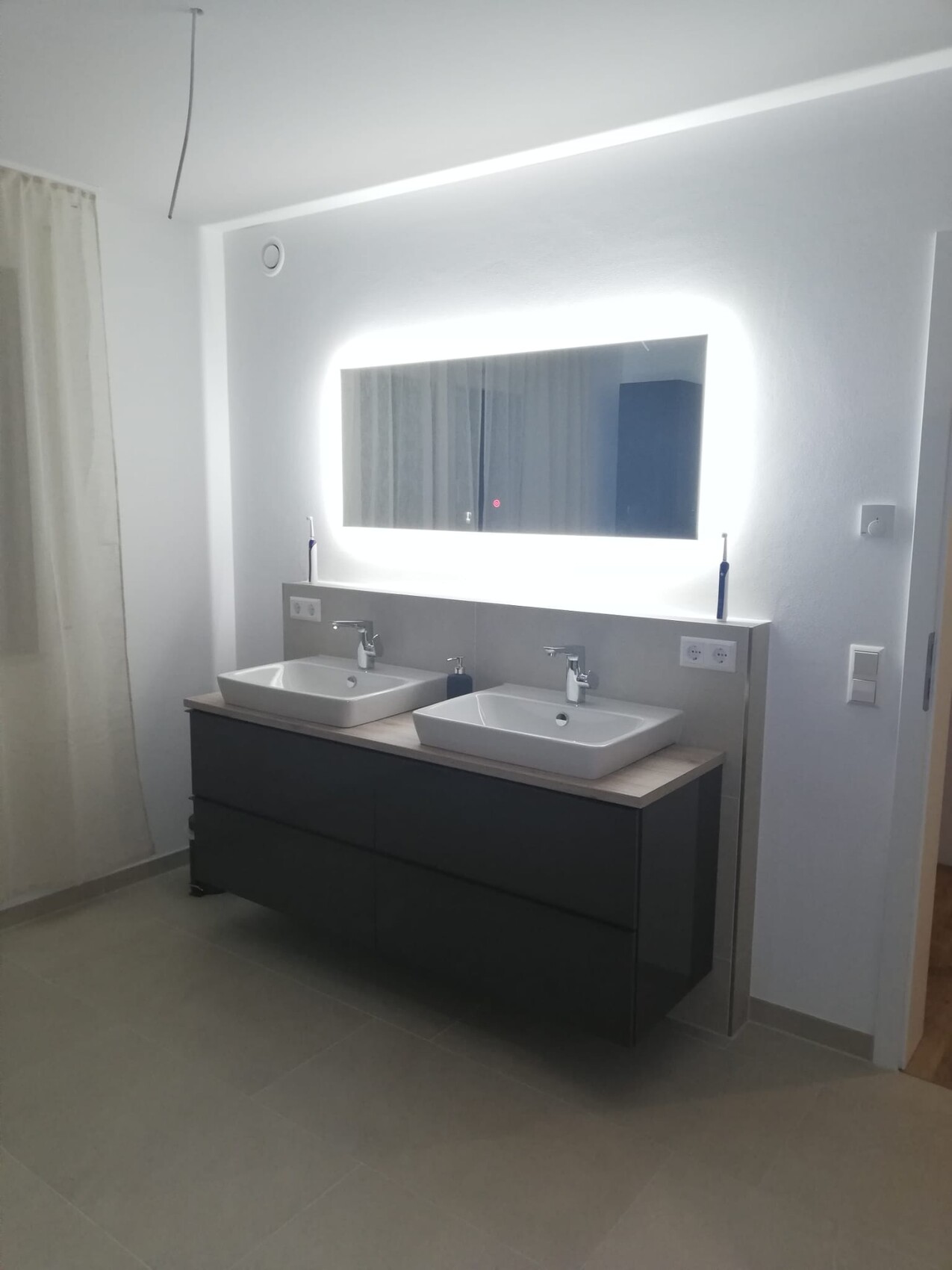 Modernes Badezimmer mit Doppelwaschbecken, dunklem Unterschrank und beleuchtetem Spiegel.