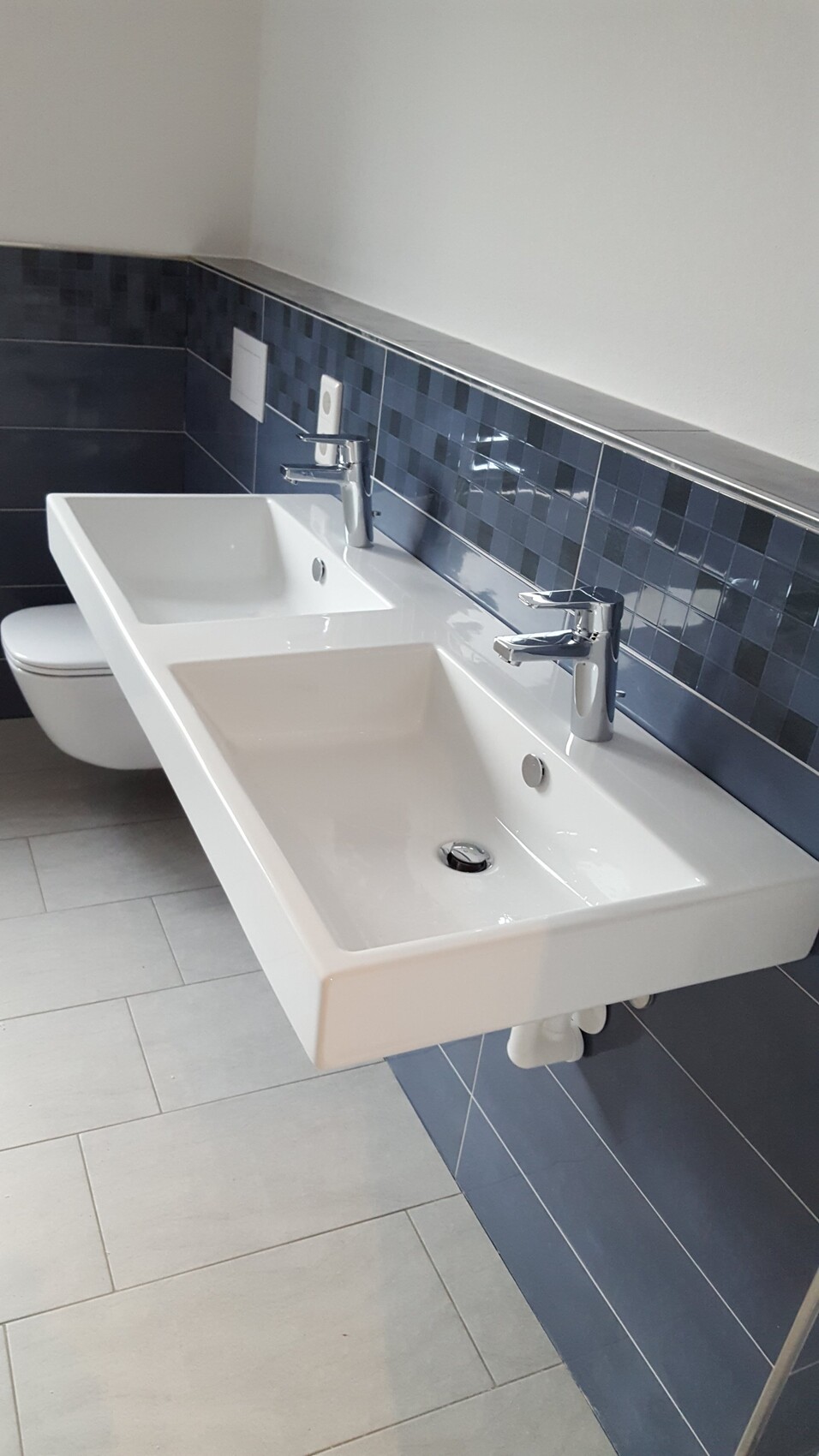 Doppel-Waschbecken mit zwei Armaturen vor blauer Mosaikfliesenwand, Toilette im Hintergrund.