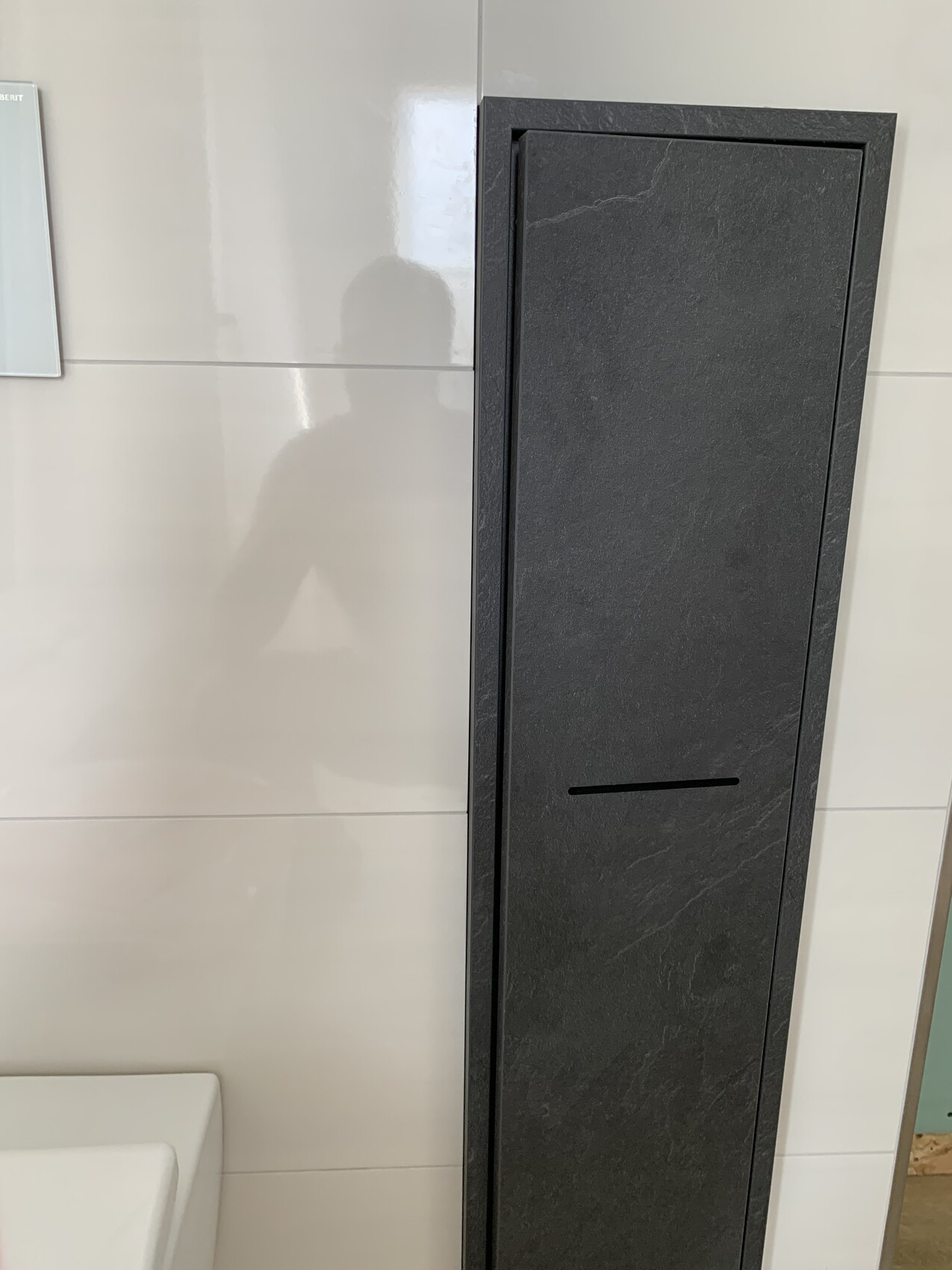 Dunkler, schmaler Wandschrank mit horizontaler Schlitzöffnung in einem hellen Badezimmer.