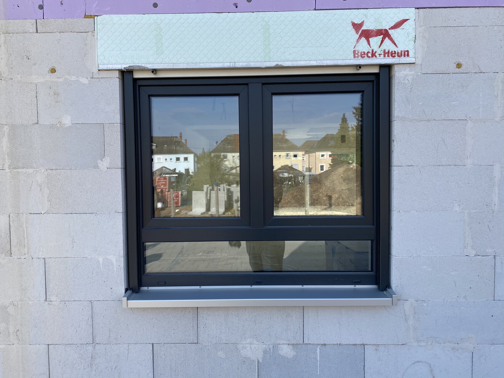 Fenster mit schwarzem Rahmen in grauer Steinwand; Baustellenreflexion zeigt Häuser.