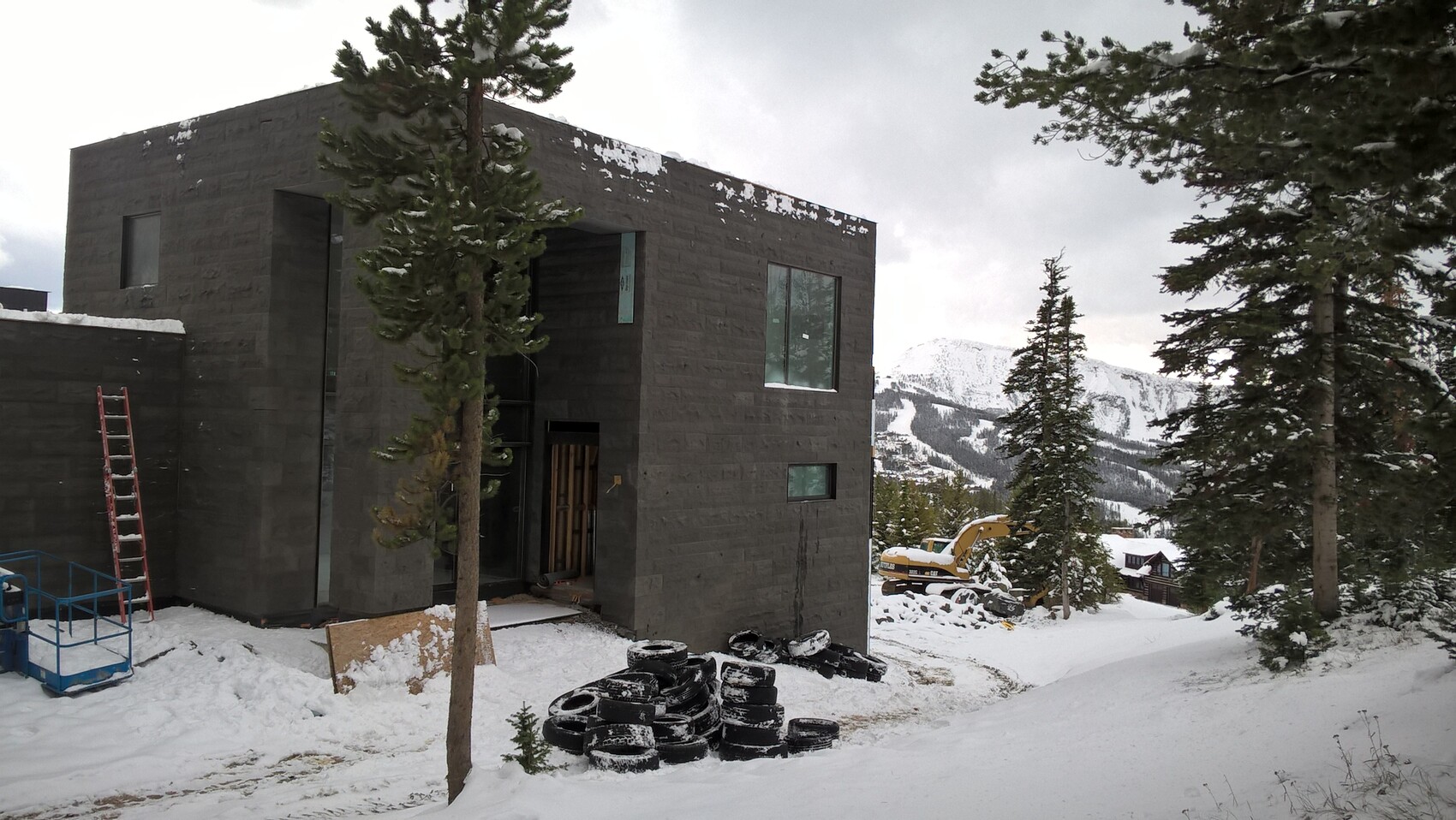 Moderne graue Steinbau-Fassade im Winter mit Schnee, Baugeräte und Reifenstapel.