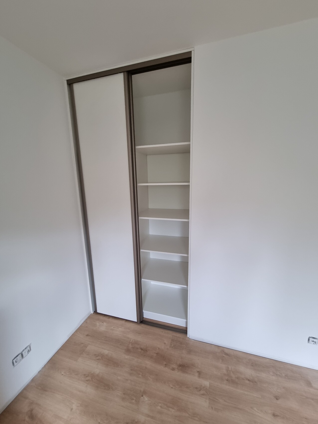 Kleiderschrank mit zwei Schiebetüren, eine Tür geöffnet, Innenregale sichtbar, heller Boden.