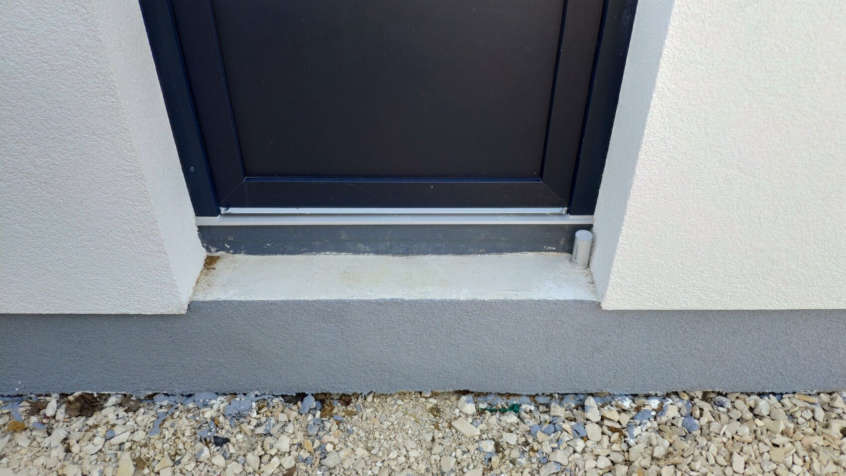 Dunkle Tür mit dunklem Rahmen vor heller Außenwand, vordere Schwelle, Kiesboden davor, rechts kleines weißes Rohr.