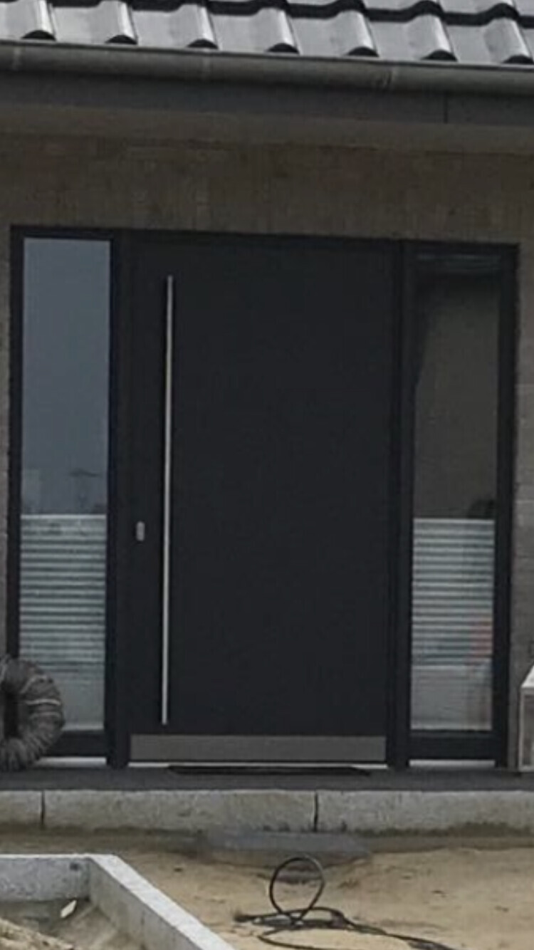Moderne schwarze Haustür mit vertikalem Griff, flankierenden Glasfenstern vor grauer Fassade.