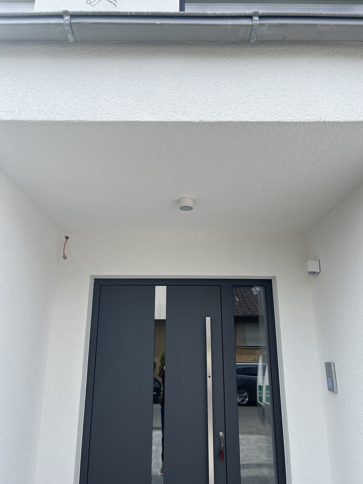 Moderne dunkle Haustür mit vertikaler Griffleiste vor weißer Putzfassade und Deckenleuchte.