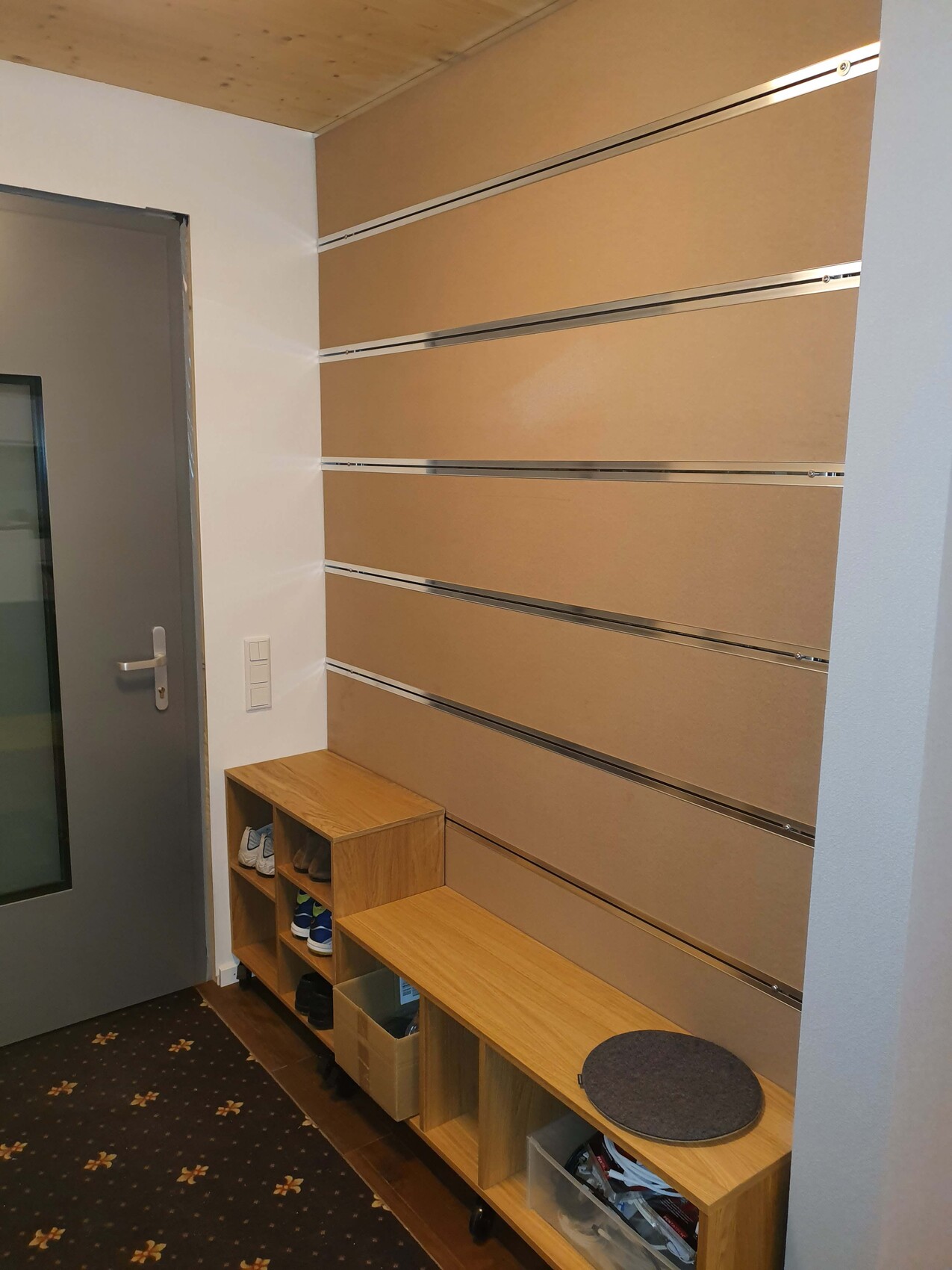 Eingangsbereich mit Holzschuhregal, Bank, runder Sitzmatte, beige Wandpaneele und grauer Tür.