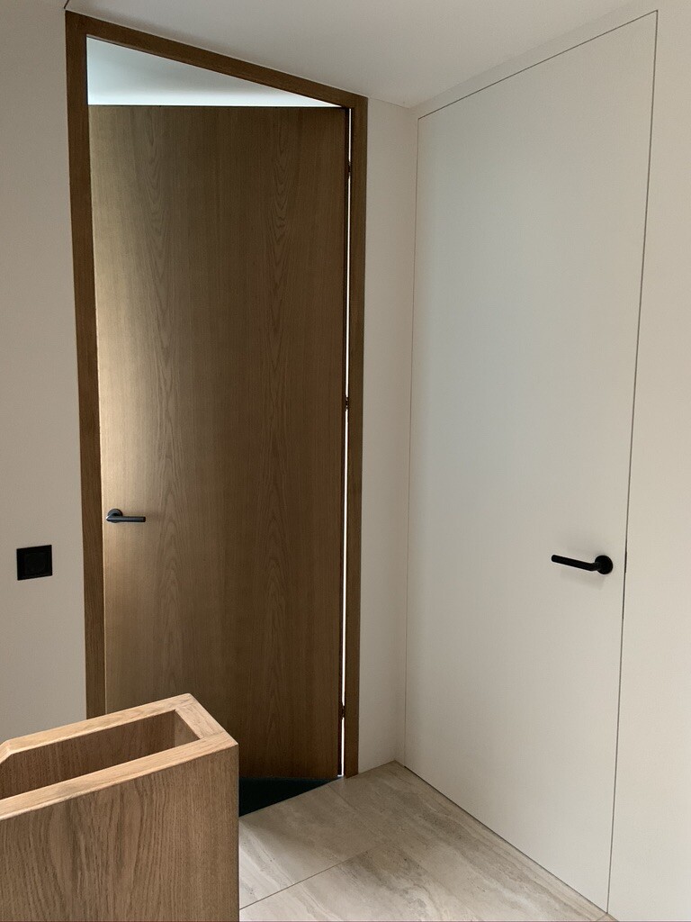 Innenansicht: zwei Türen (braun links, weiß rechts), heller Fliesenboden, Holzkasten im Vordergrund.